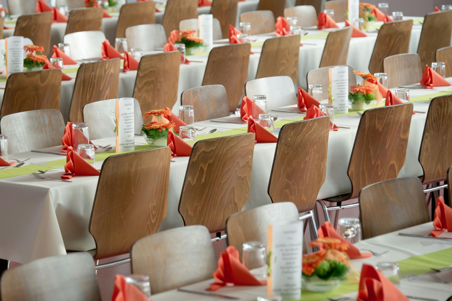 Rows of tables set for a banquet; white tablecloths, wood-backed chairs, orange napkins, and floral centerpieces.
