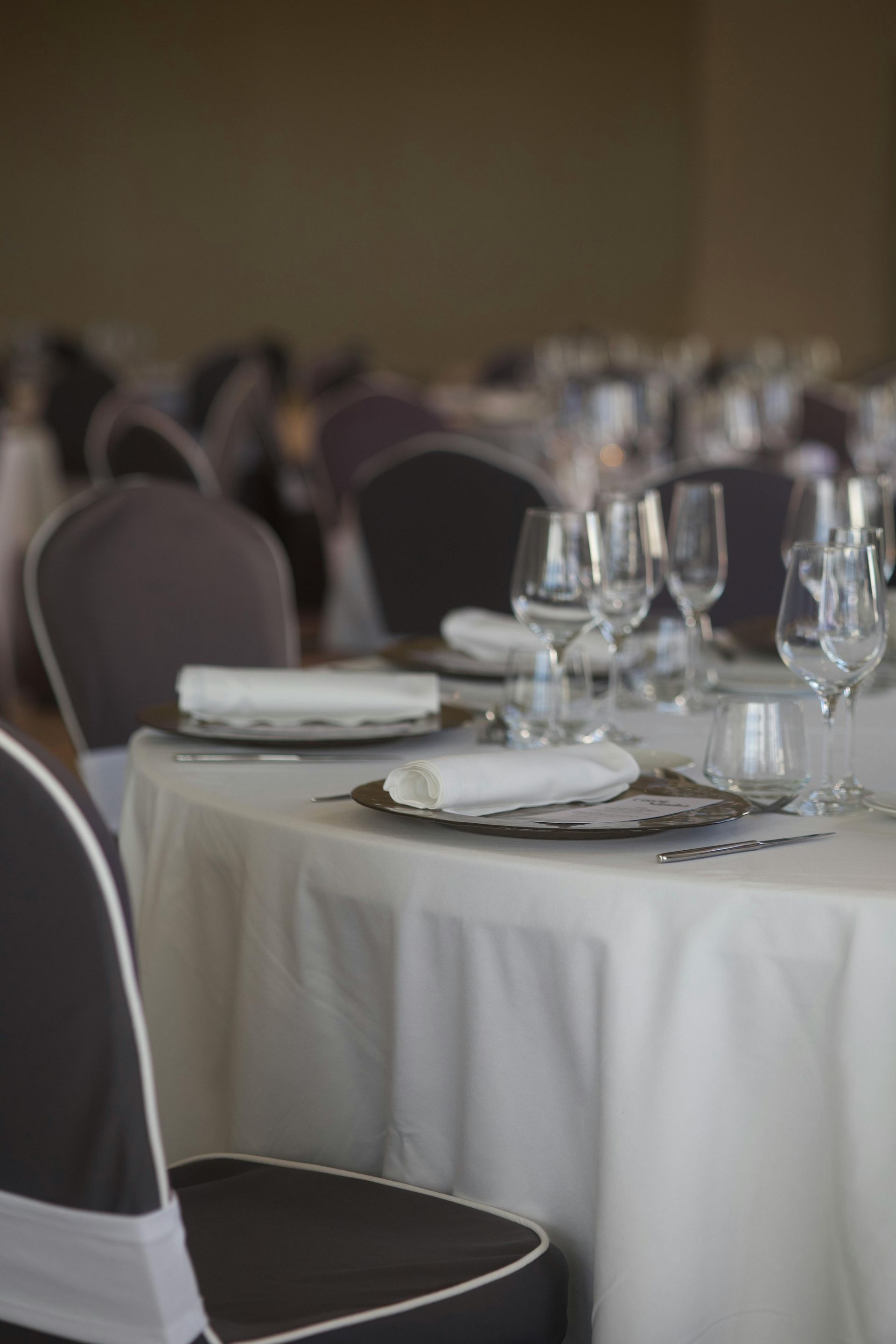 Table setting in a banquet hall: white tablecloth, plates, glasses, and chairs with gray covers.