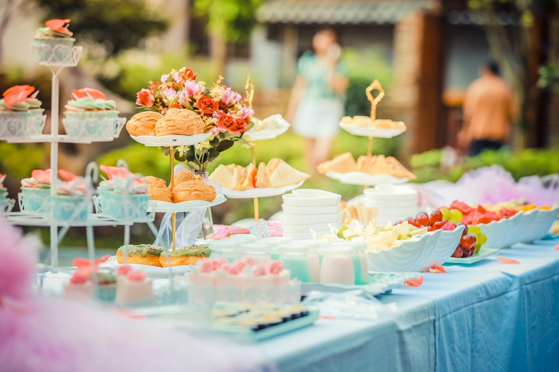 Dessert table with tiered treats and flowers; party in garden setting.