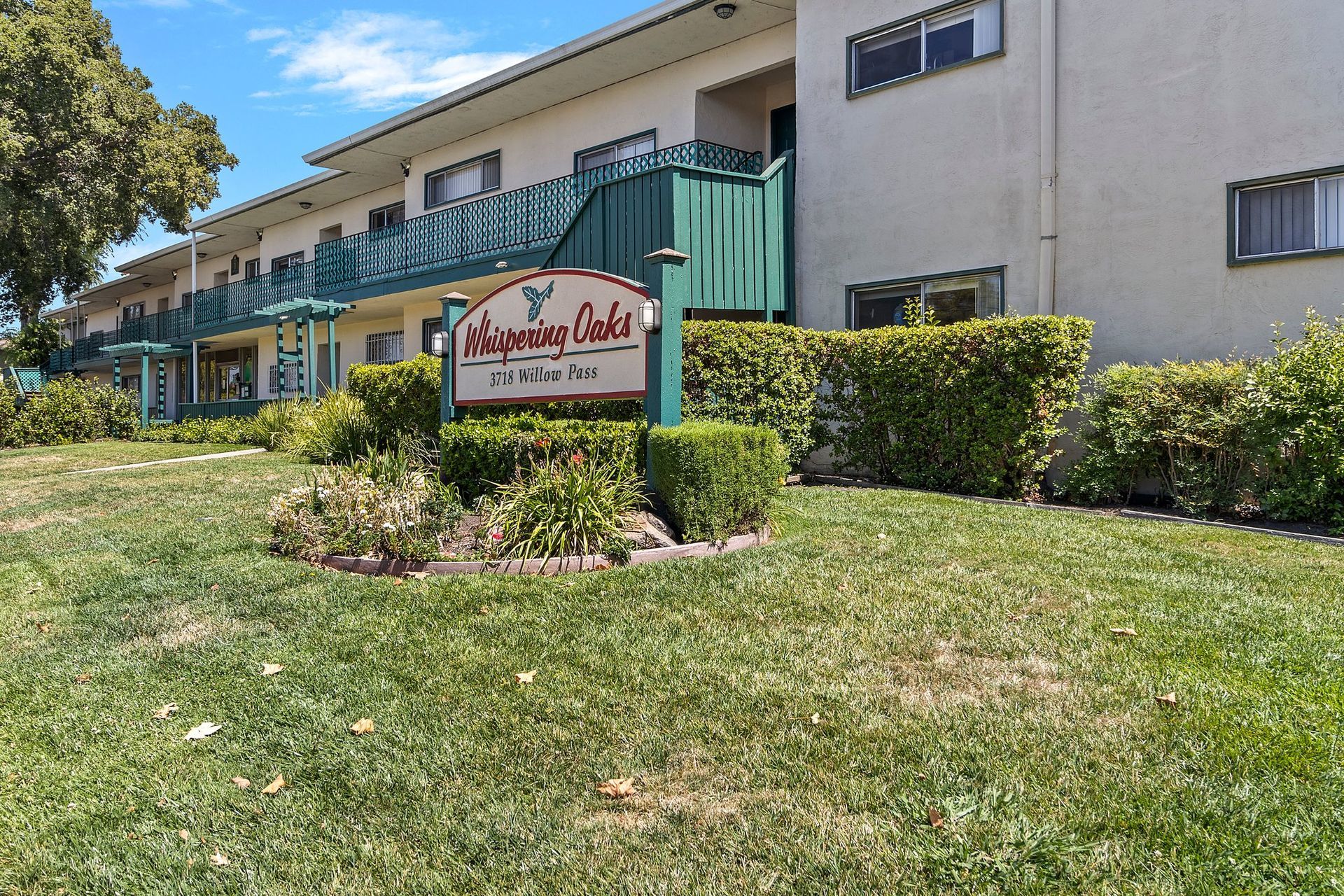A low-angle view of a beige apartment building with green balconies and a Whispering Oaks sign on a grassy lawn at Whispering Oaks in Concord, CA.