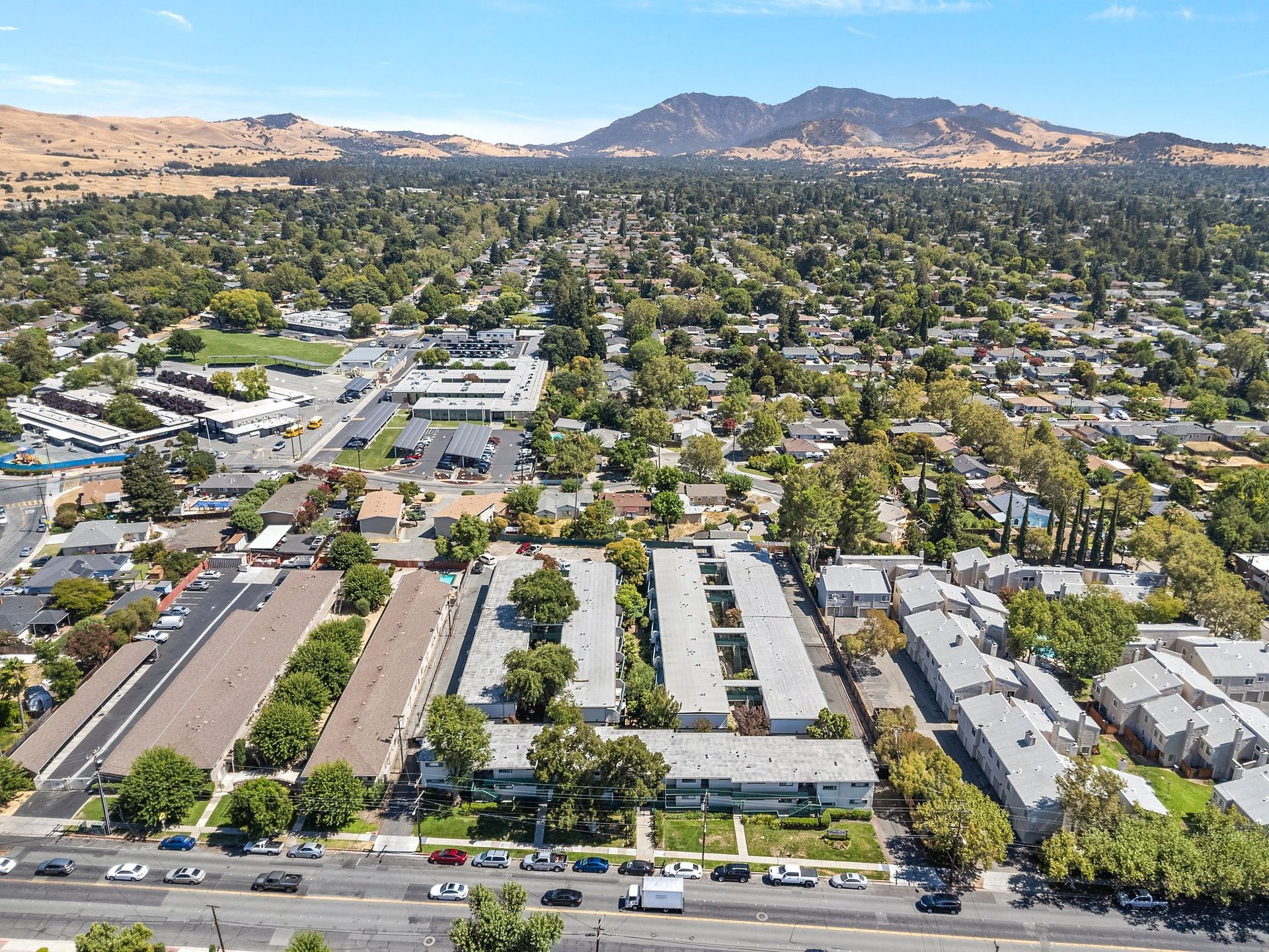 Aerial view of an apartment complex near a busy road, with residential neighborhoods and distant mountains in the background.
