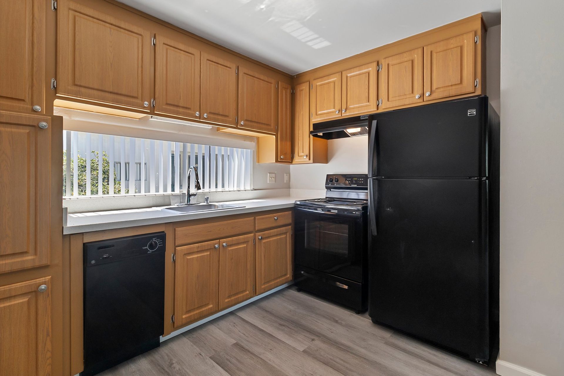 A small kitchen with light wood cabinets, light-toned wood-look flooring, and black appliances including a fridge and stove at Whispering Oaks in Concord, CA.