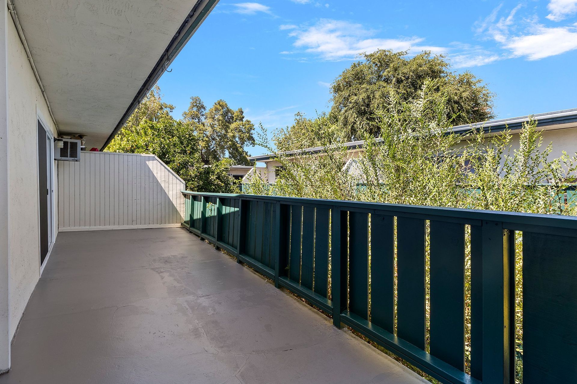A balcony with a grey floor and a green wooden railing, overlooking trees and buildings under a sunny blue sky at Whispering Oaks in Concord, CA.