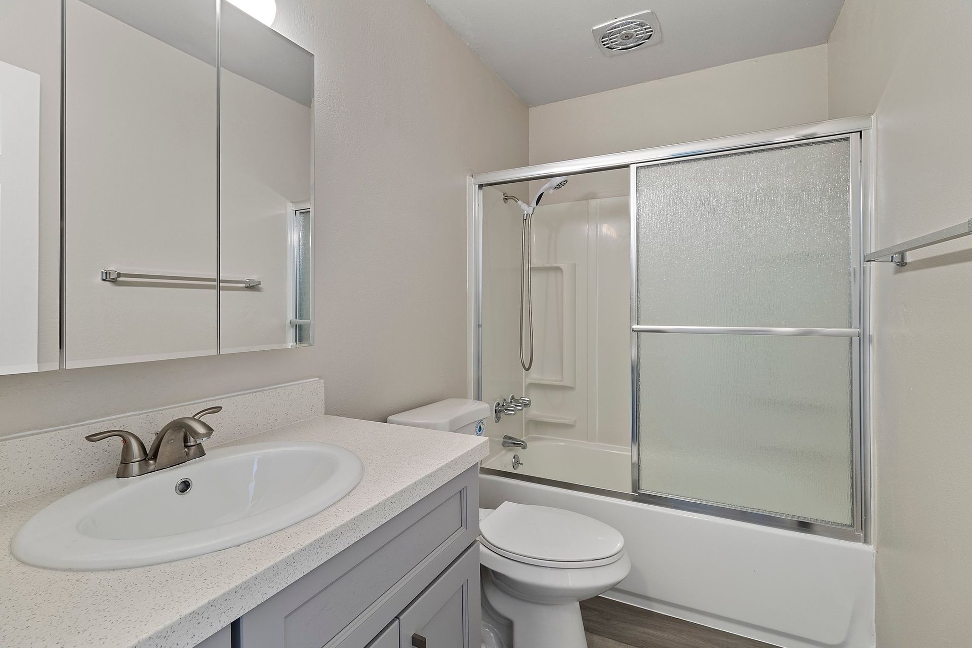 A modern bathroom with a light grey vanity, white countertop, sink, mirror, toilet, and a glass-enclosed bathtub shower at Whispering Oaks in Concord, CA.