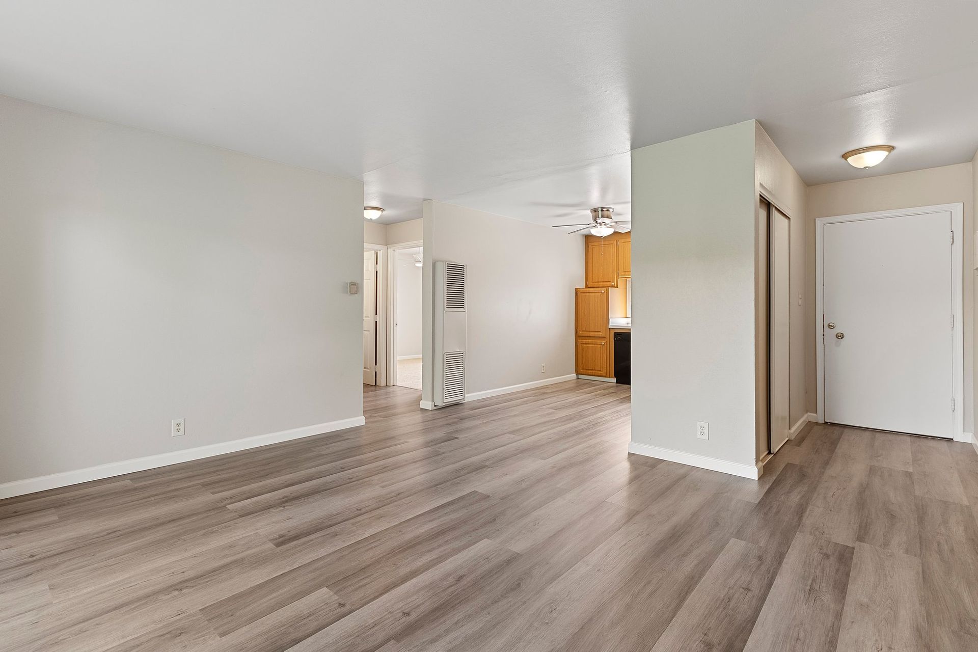 An empty room with light gray walls and wood-patterned flooring, showing an open doorway and a kitchen area in the back at Whispering Oaks in Concord, CA.
