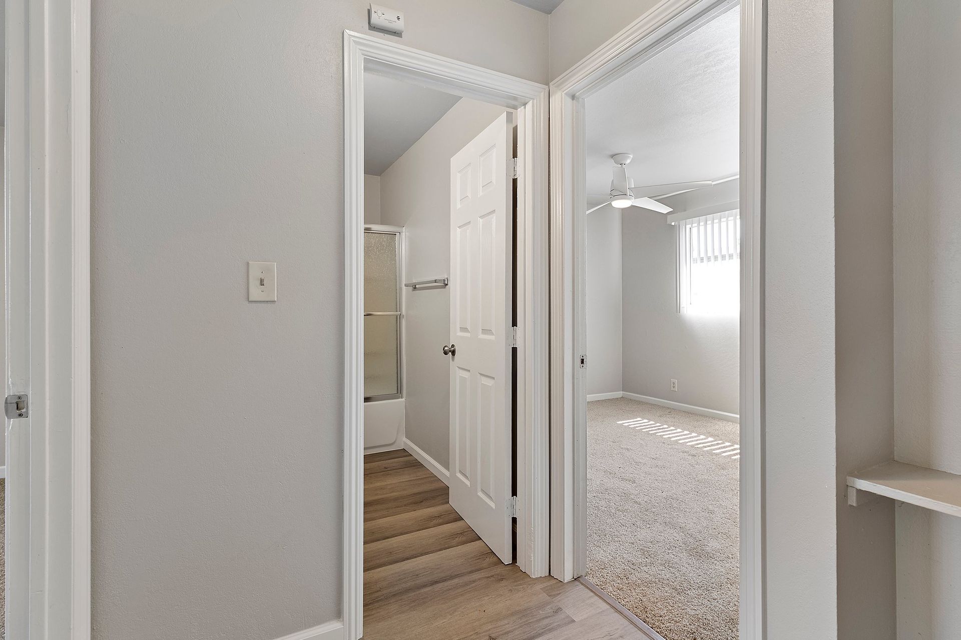 A bright indoor hallway showing two doorways: one leading to a bathroom with wood-look flooring, the other to a carpeted room at Whispering Oaks in Concord, CA.