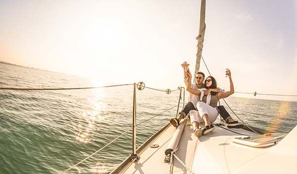 Couple on sailboat, arms raised in celebration against a sunny seascape.