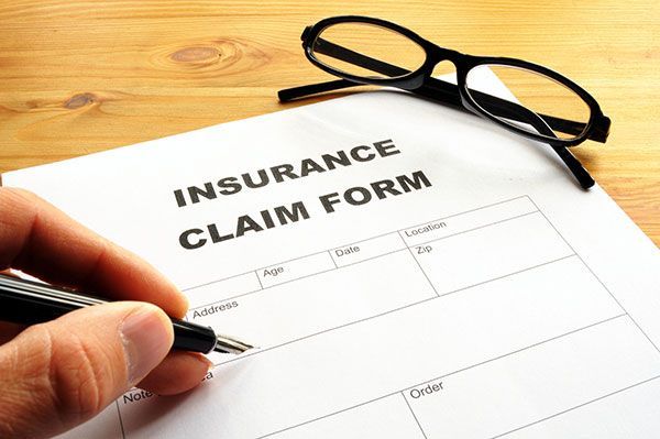 A hand filling out an insurance claim form, glasses on top, on a wooden surface.