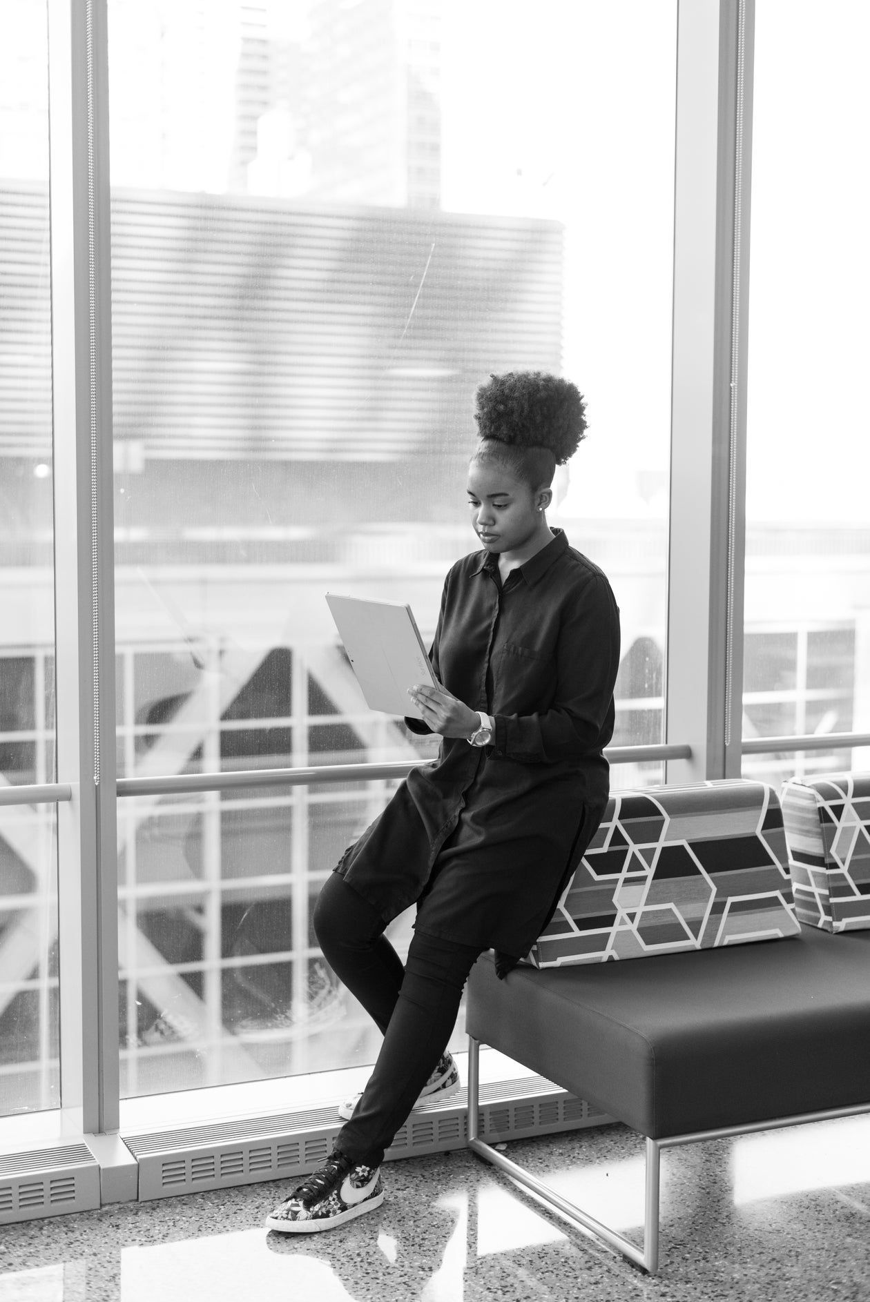 Person seated on a bench by large windows, looking at a phone in a bright lounge area.