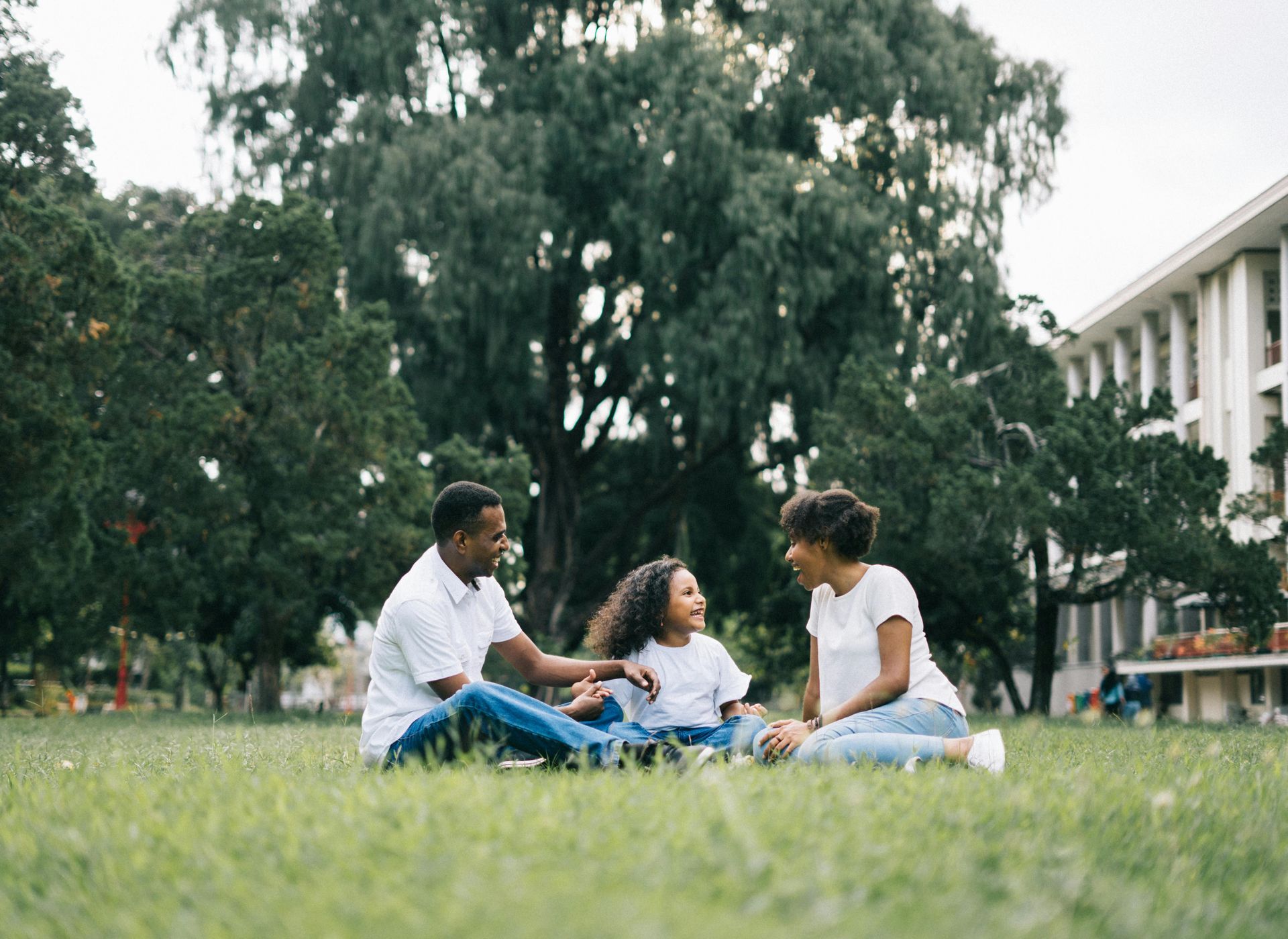 Three people sitting on grass in a park, talking and smiling near trees and a building.