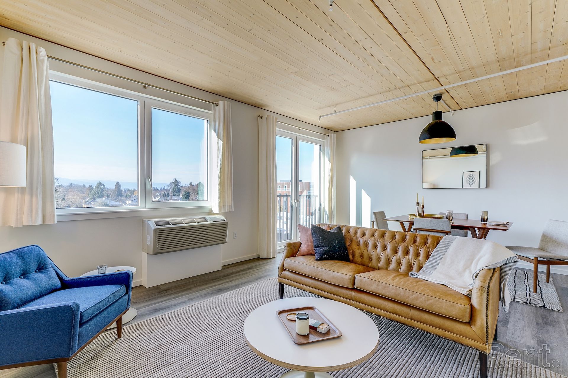 Living room with large windows, blue armchair, tan leather sofa, dining table, and round coffee table.