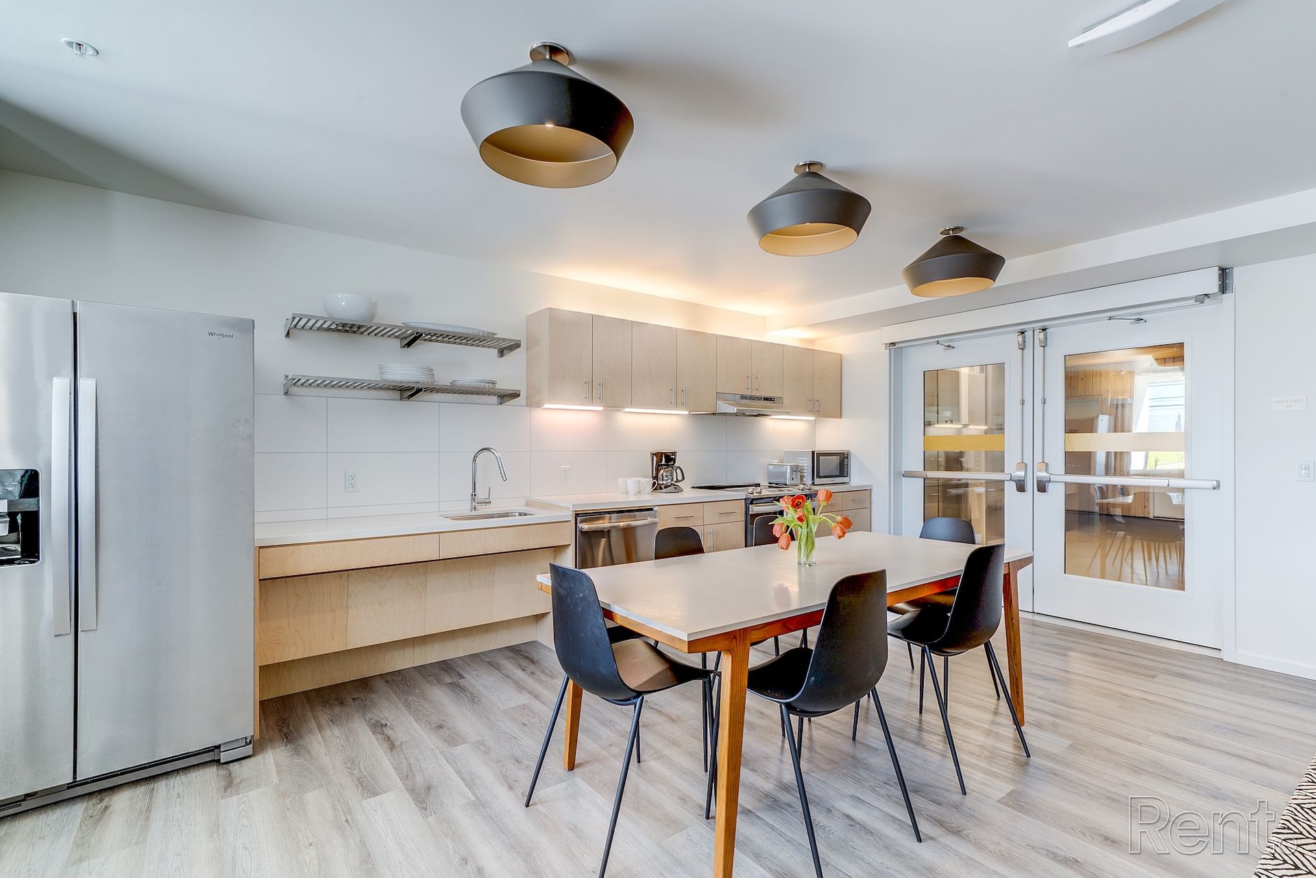 Modern kitchen/dining area with light wood cabinets, stainless steel appliances, and a table with black chairs.
