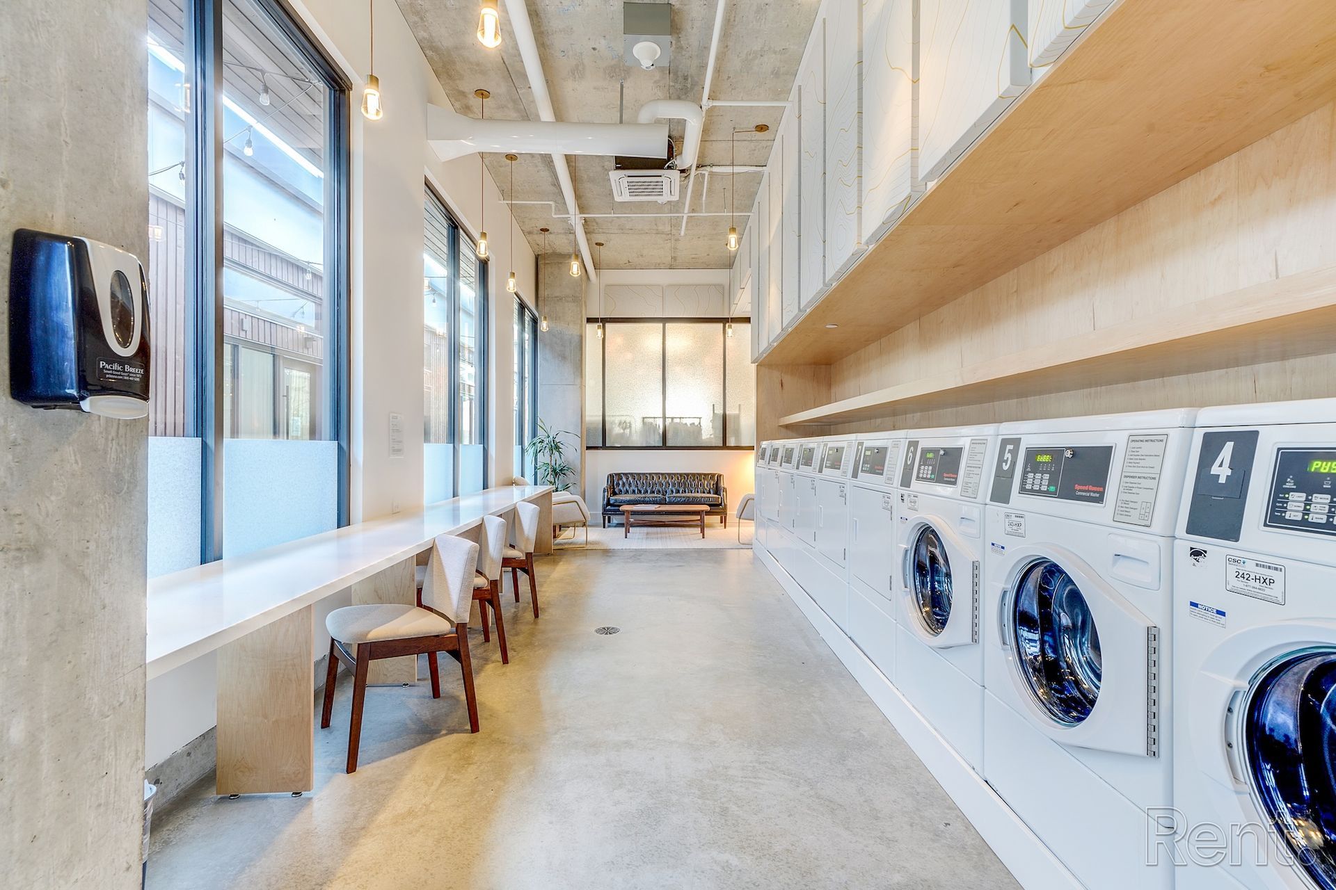 Laundry room with rows of washing machines, seating area, and large windows.