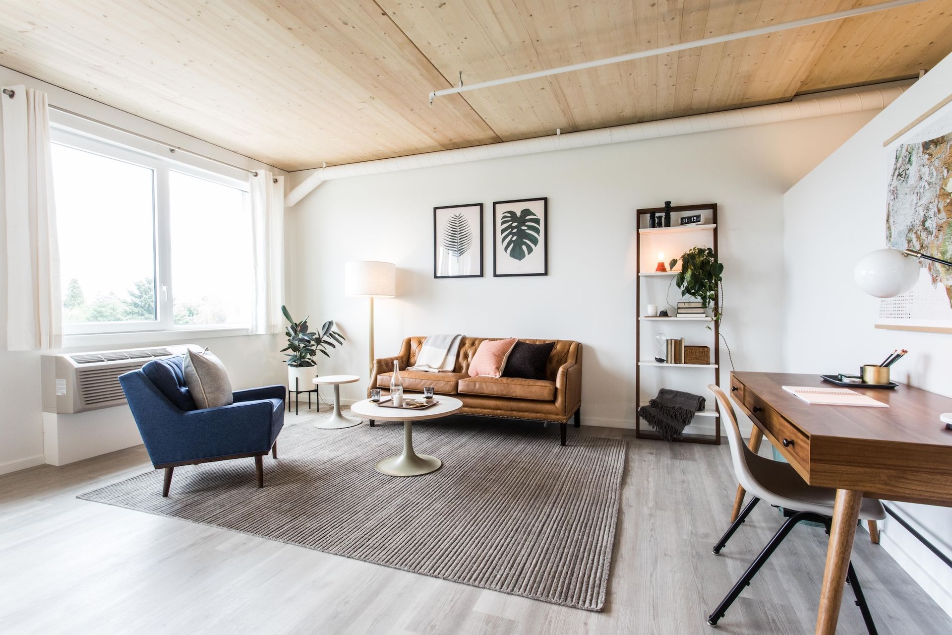 Living room with blue armchair, brown sofa, and wooden desk. Natural light and neutral colors.