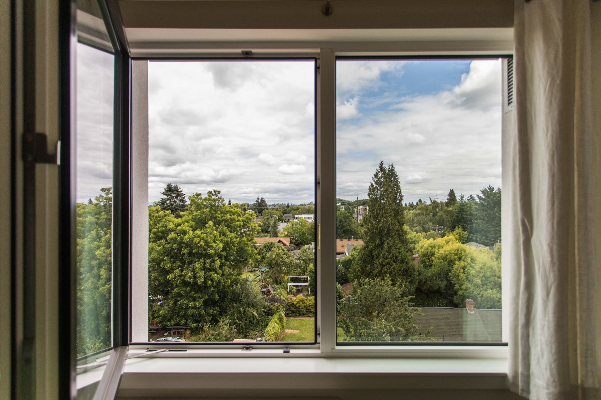Open window with view of trees, houses, and cloudy sky. White window frame, white curtains.