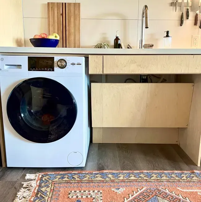 A white washing machine beside a light wood cabinet with a sink, cutting board, and fruit on the countertop.