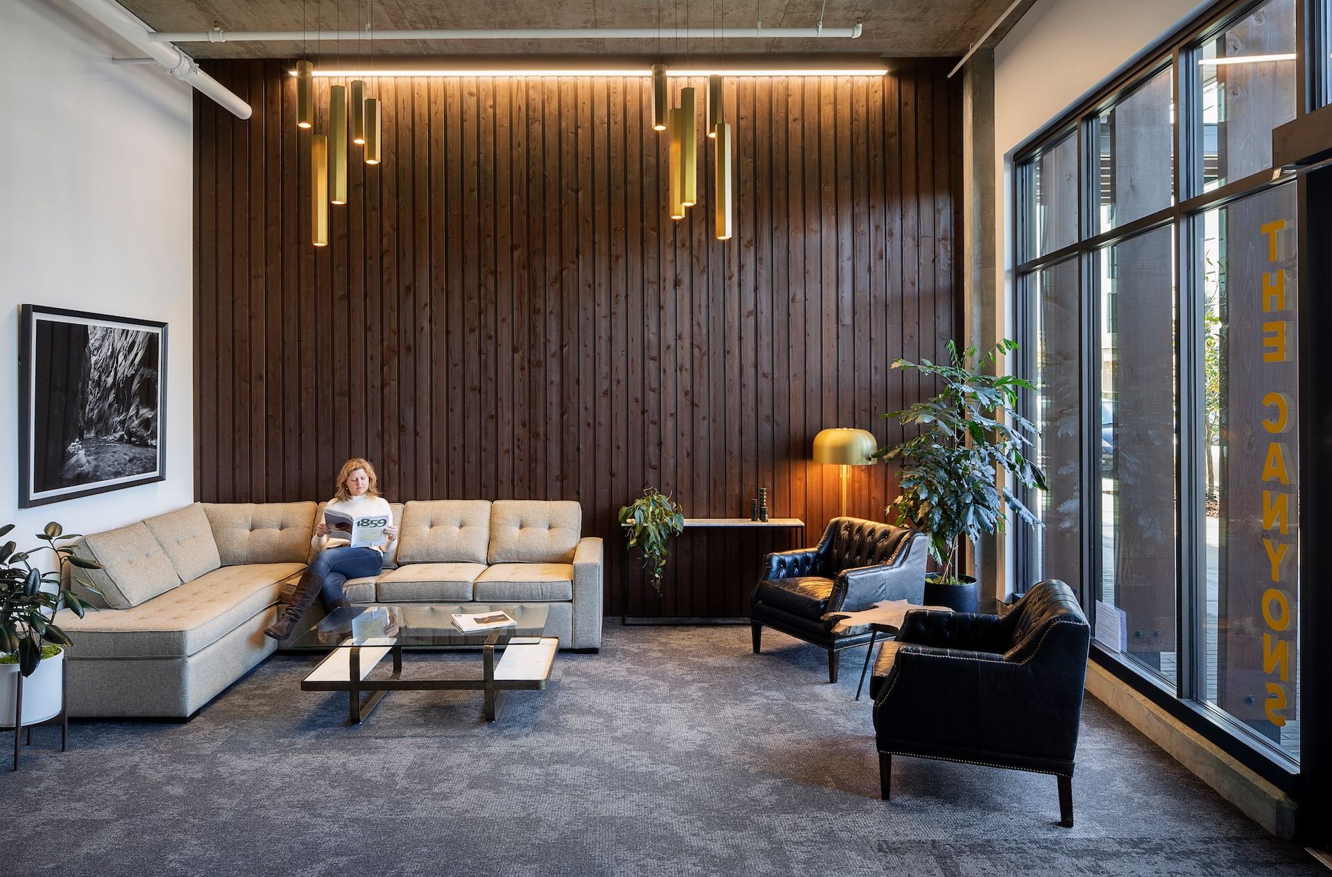 Woman seated on a sectional sofa in a modern lobby, reading. Wooden panel wall, gold lights, black armchairs, and large windows.