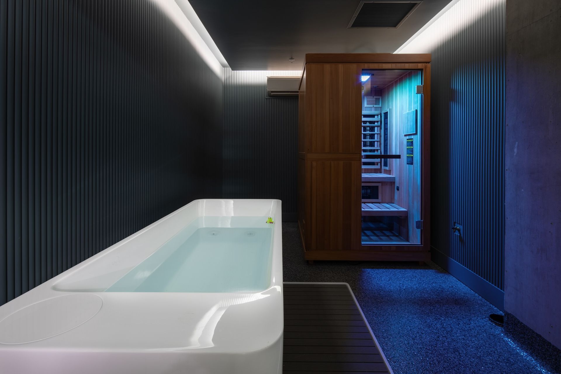 Modern spa room with a white soaking tub, dark walls, and a lit sauna at the far end