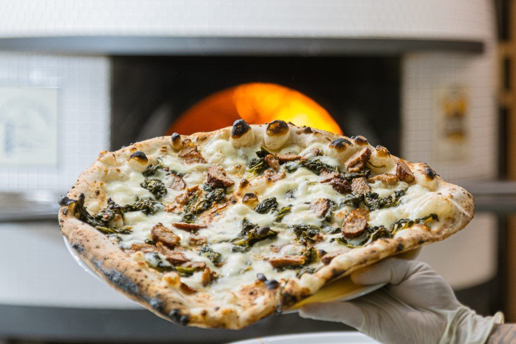 Mano che tiene una pizza condita, davanti a un forno a legna.