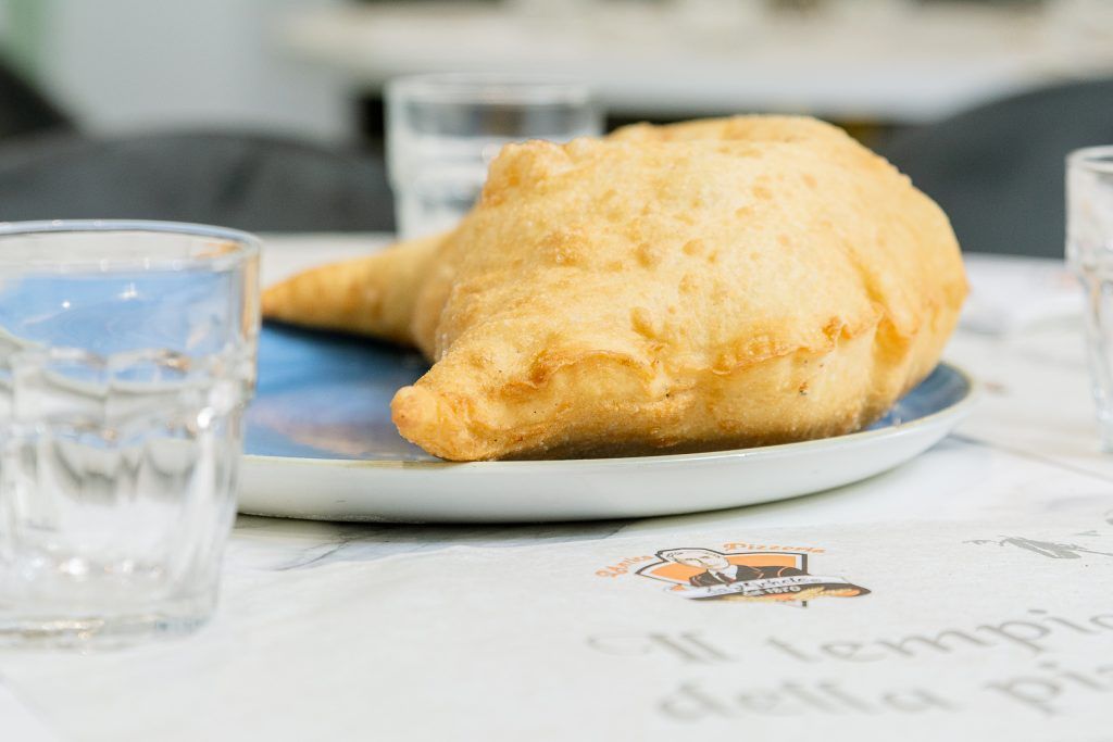 Pasta sfoglia fritta su un piatto blu, con bicchieri d'acqua su un tavolo coperto da una tovaglia fantasia.