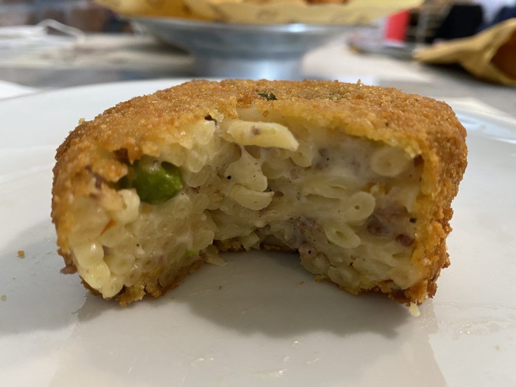 Crocchetta di pasta fritta, tagliata a metà. Presenta maccheroni, piselli e ripieno cremoso, avvolti in una croccante panatura dorata, su un piatto bianco.
