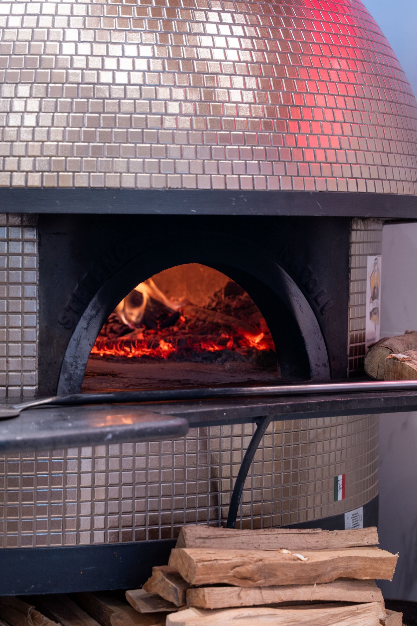 Forno a legna per pizza con fiamme e legna accatastata; esterno in piastrelle metalliche.