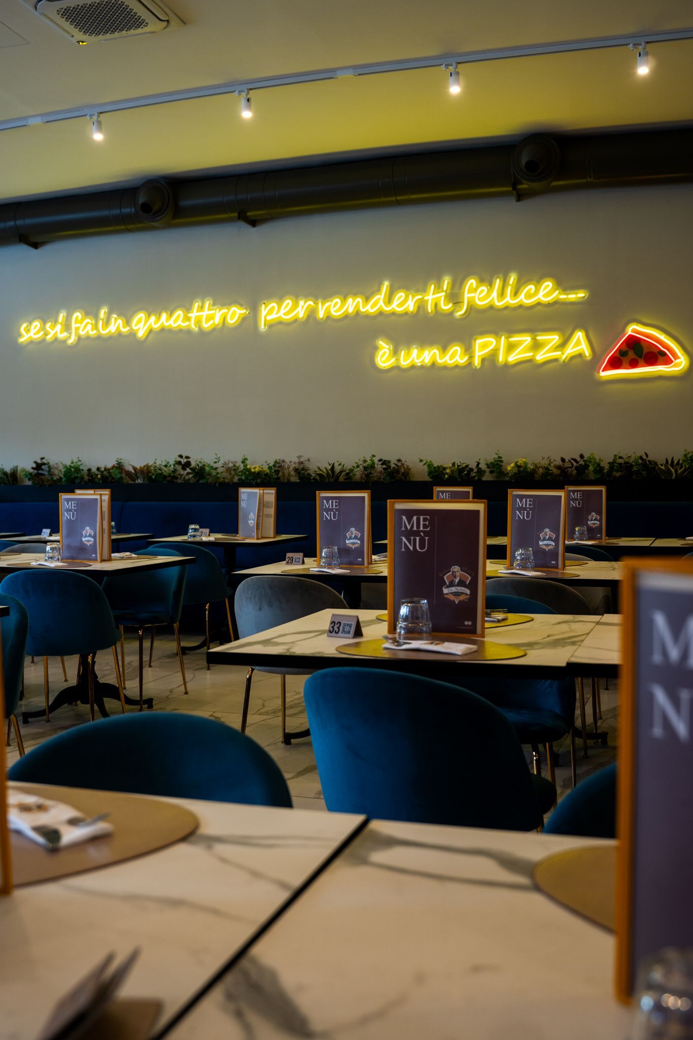 Interno di una pizzeria con tavoli, sedie blu e un'insegna al neon sul muro.