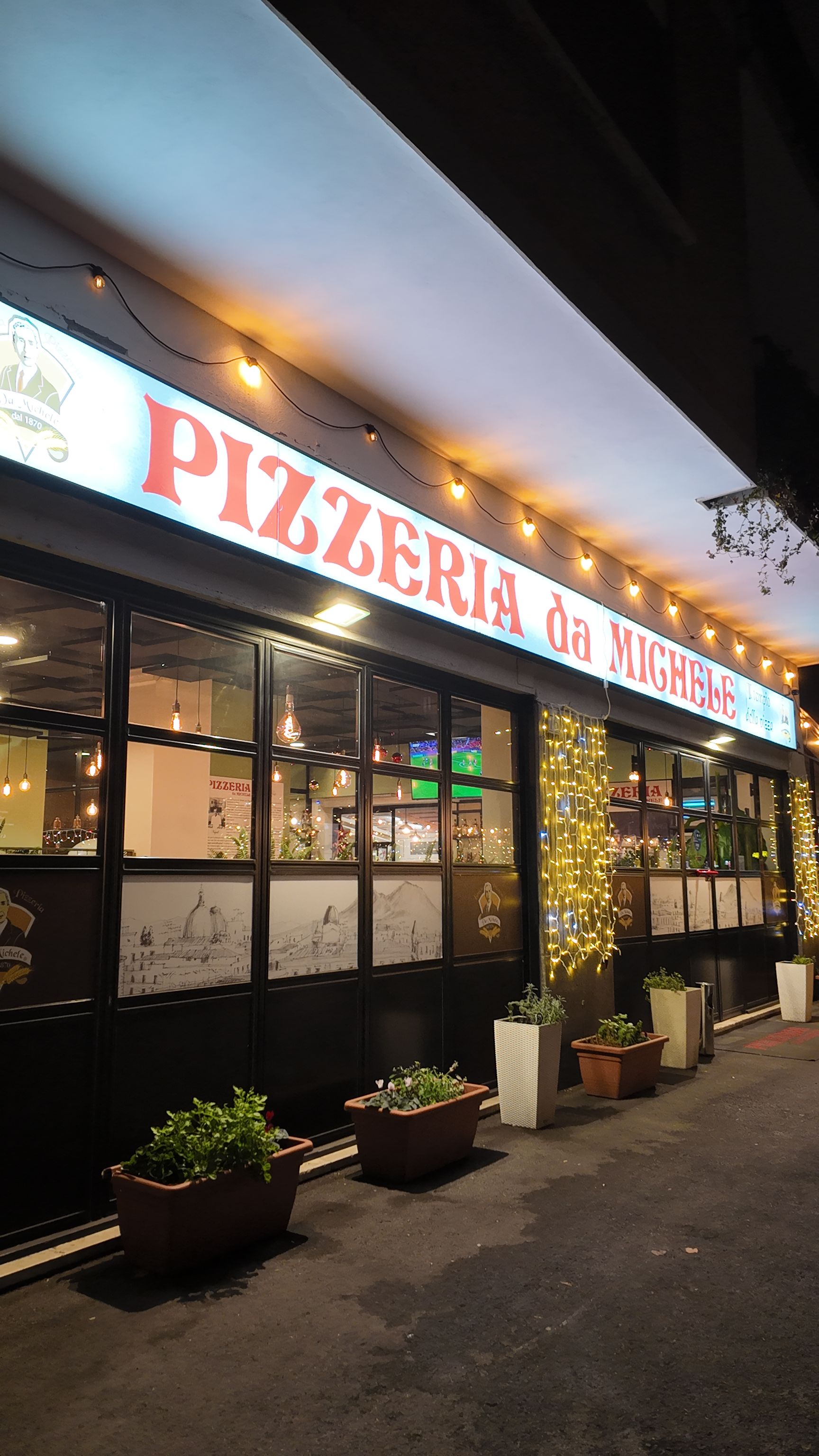 Ingresso della pizzeria di notte, insegne luminose, luci a ghirlanda. Fioriere marroni con verde sotto le finestre.