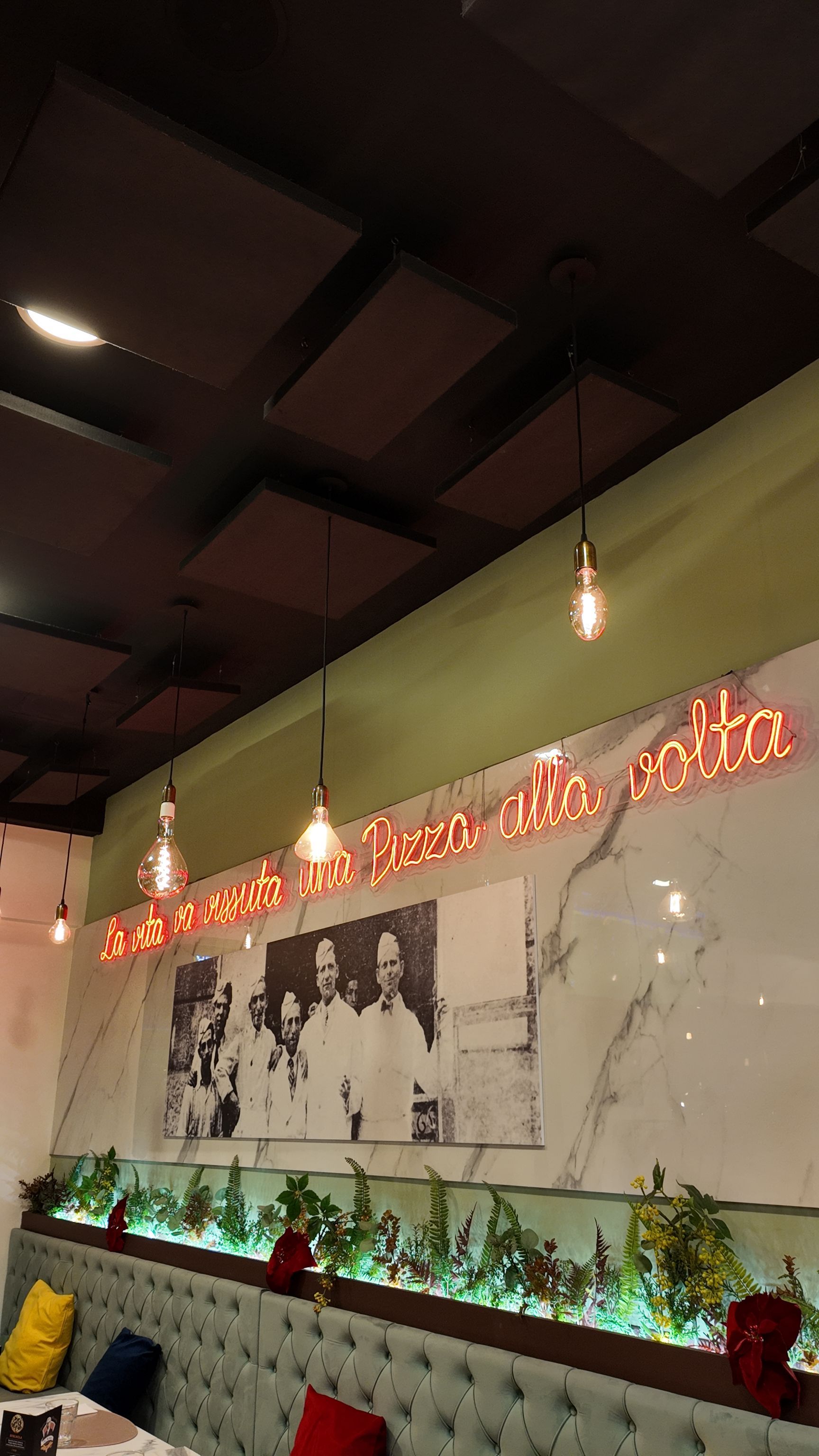 Interno del ristorante con insegna al neon, luci sospese, una foto in bianco e nero e piante.