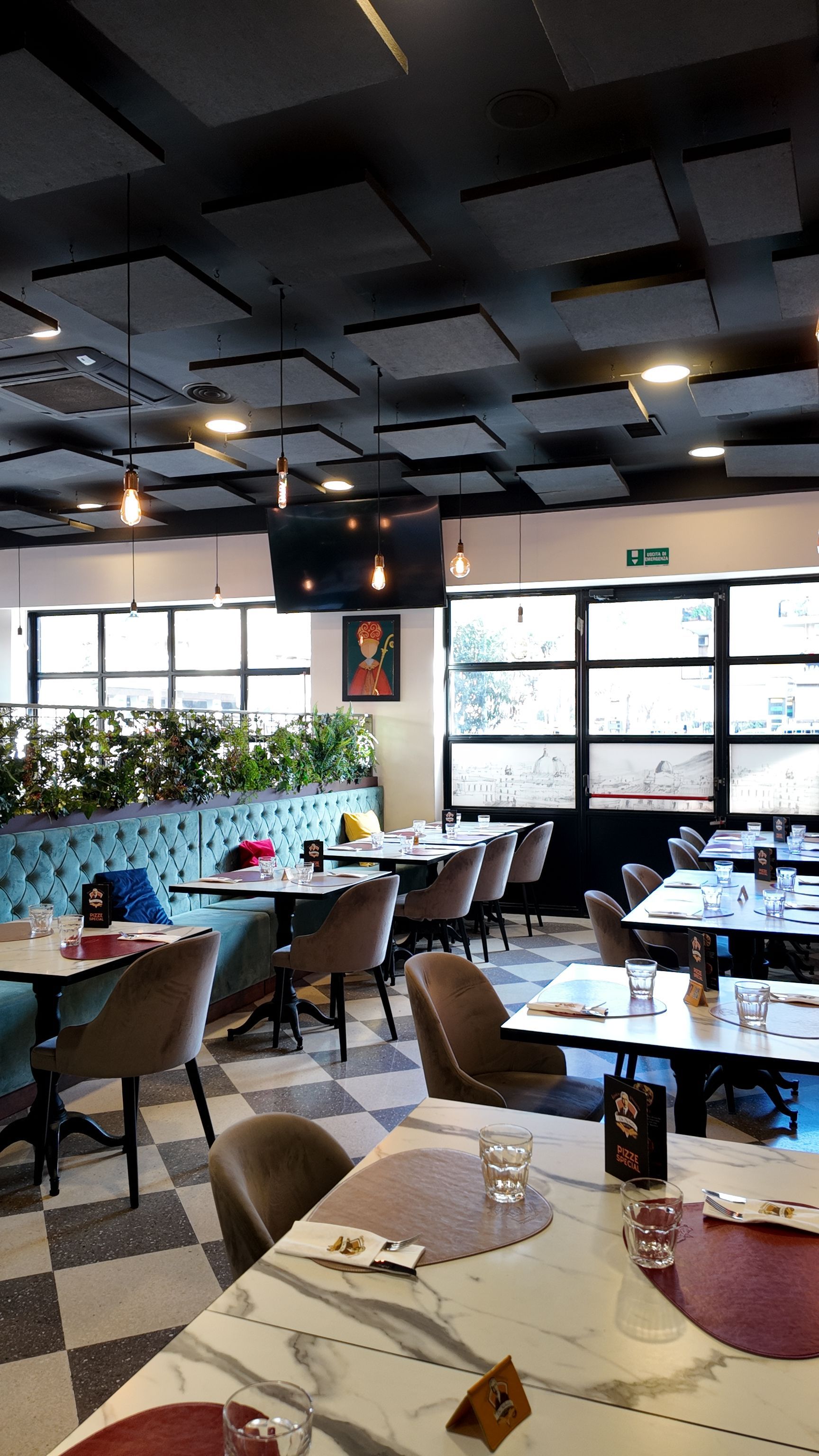 Interno del ristorante con tavoli, sedie e separé. Piante verdi, finestre e pannelli scuri sul soffitto.
