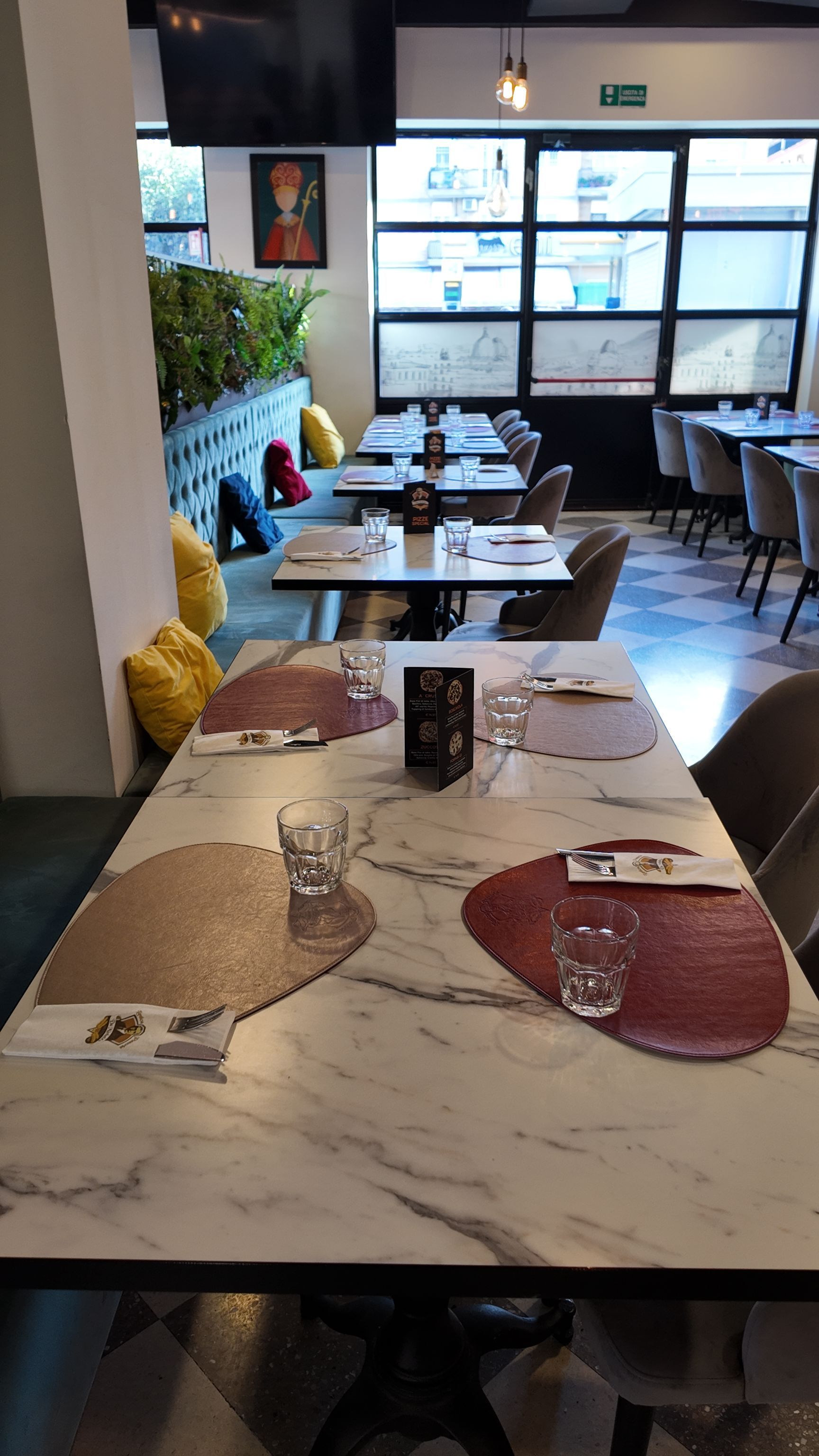 Interno del ristorante: tavoli apparecchiati per la cena, con tovagliette decorative e bicchieri. Finestre sullo sfondo.