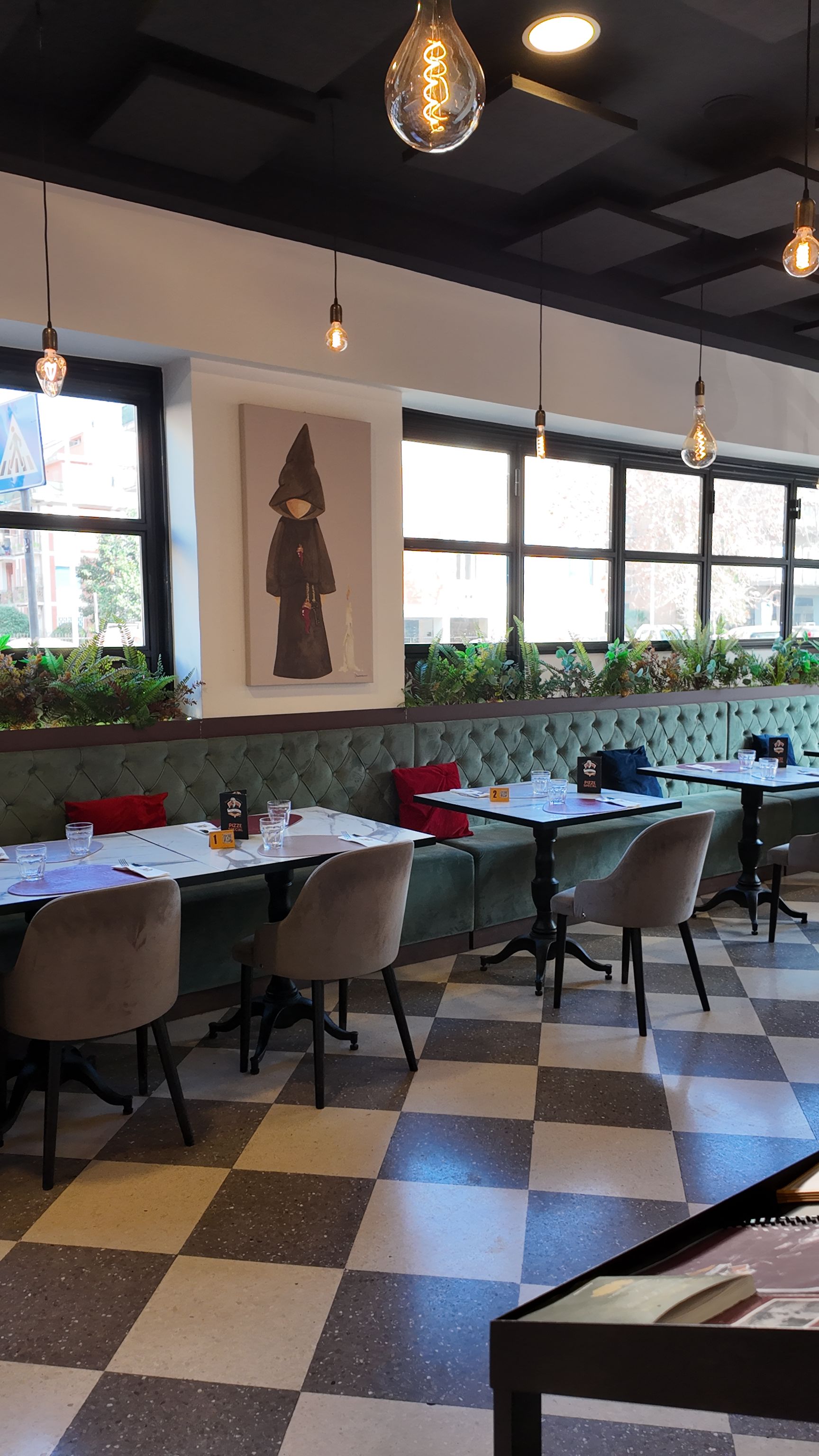 Interno del ristorante con pavimento a scacchi, tavoli, separé con cuscini verdi, ampie finestre e lampade a sospensione.