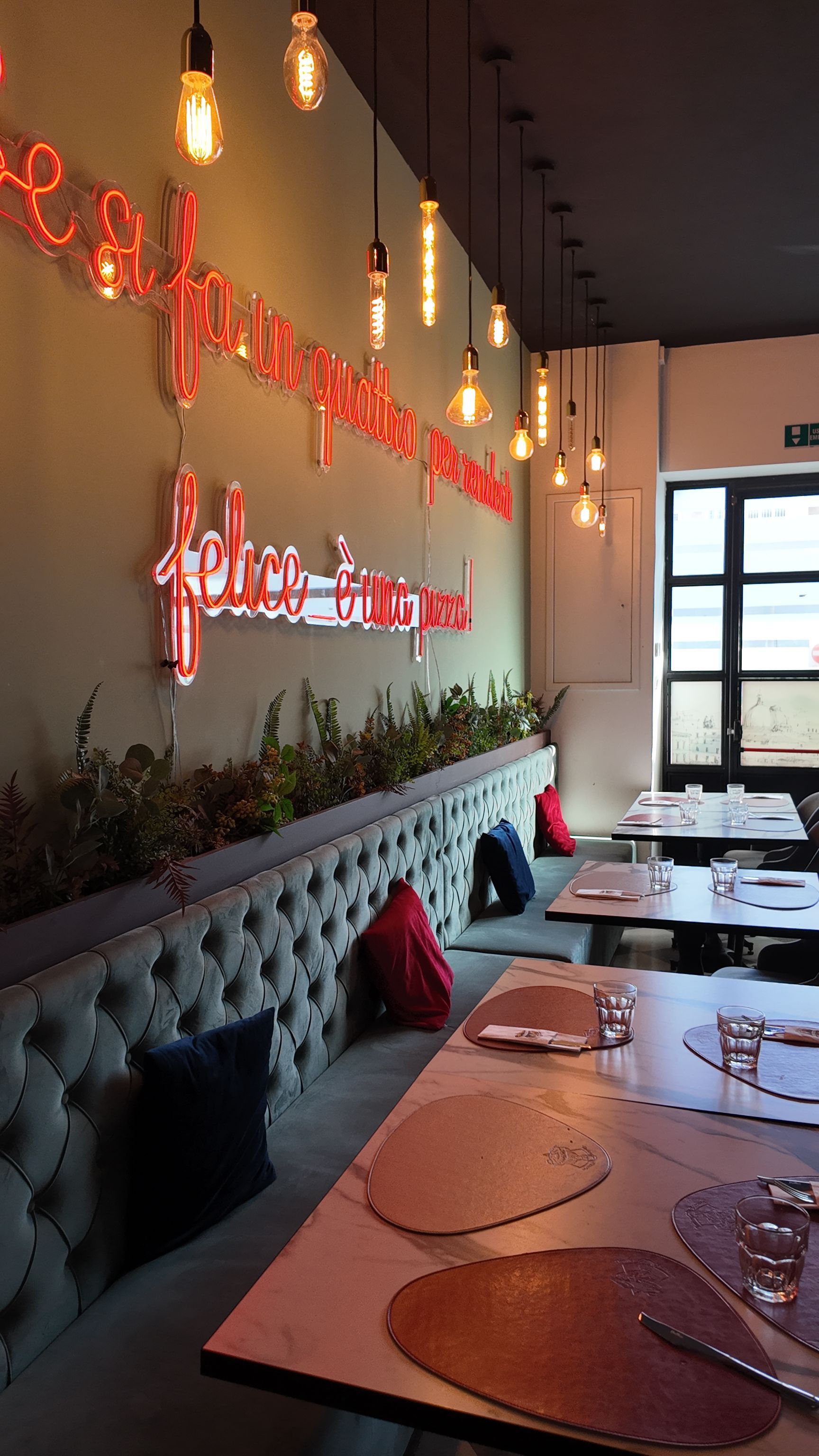 Interno del ristorante con sedute a panca, tavoli apparecchiati con tovagliette all'americana e luci sospese. Insegna al neon sulla parete.