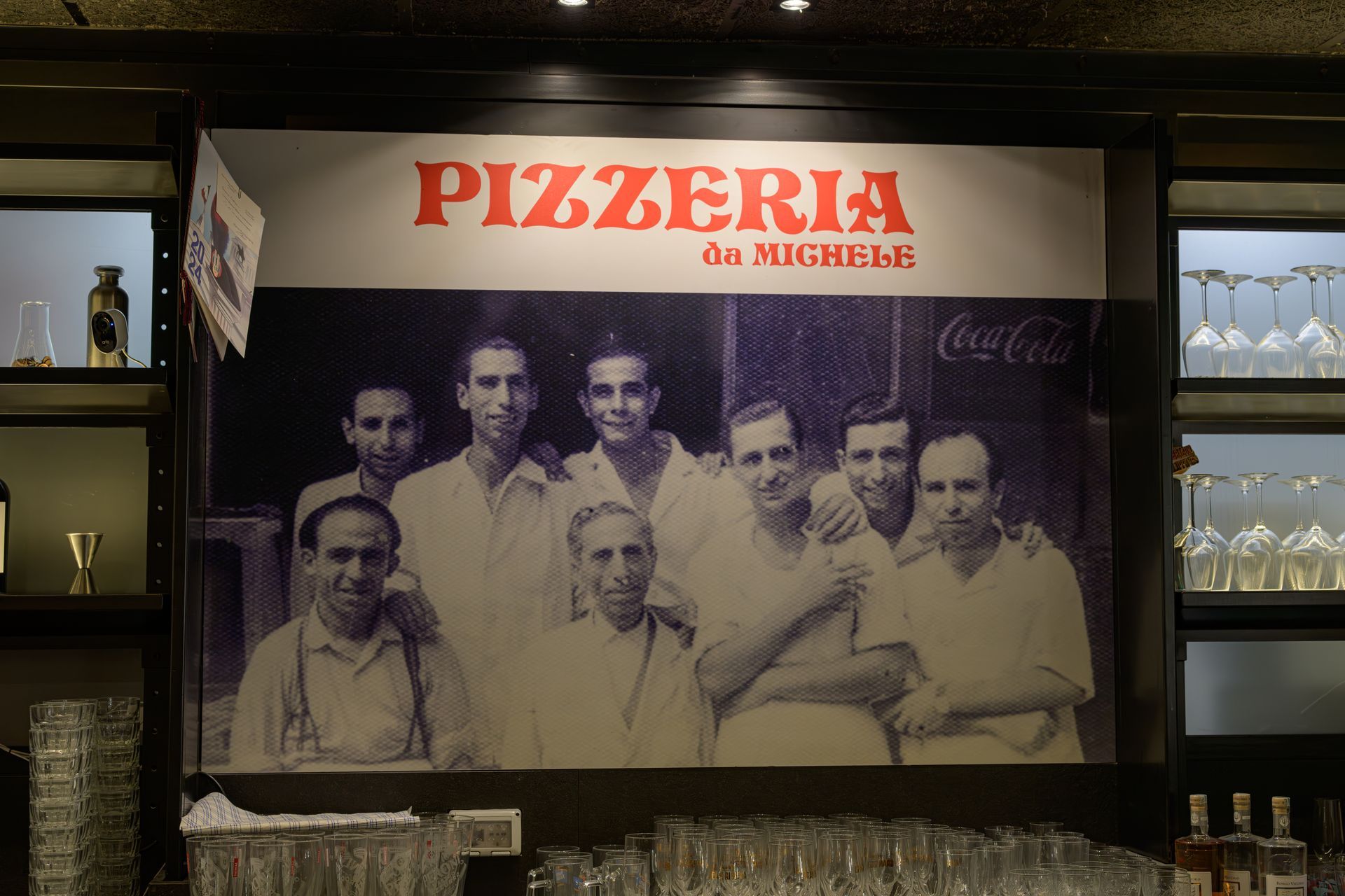 Insegna di pizzeria con una foto d'epoca di uomini; bicchieri e bottiglie visibili sotto.