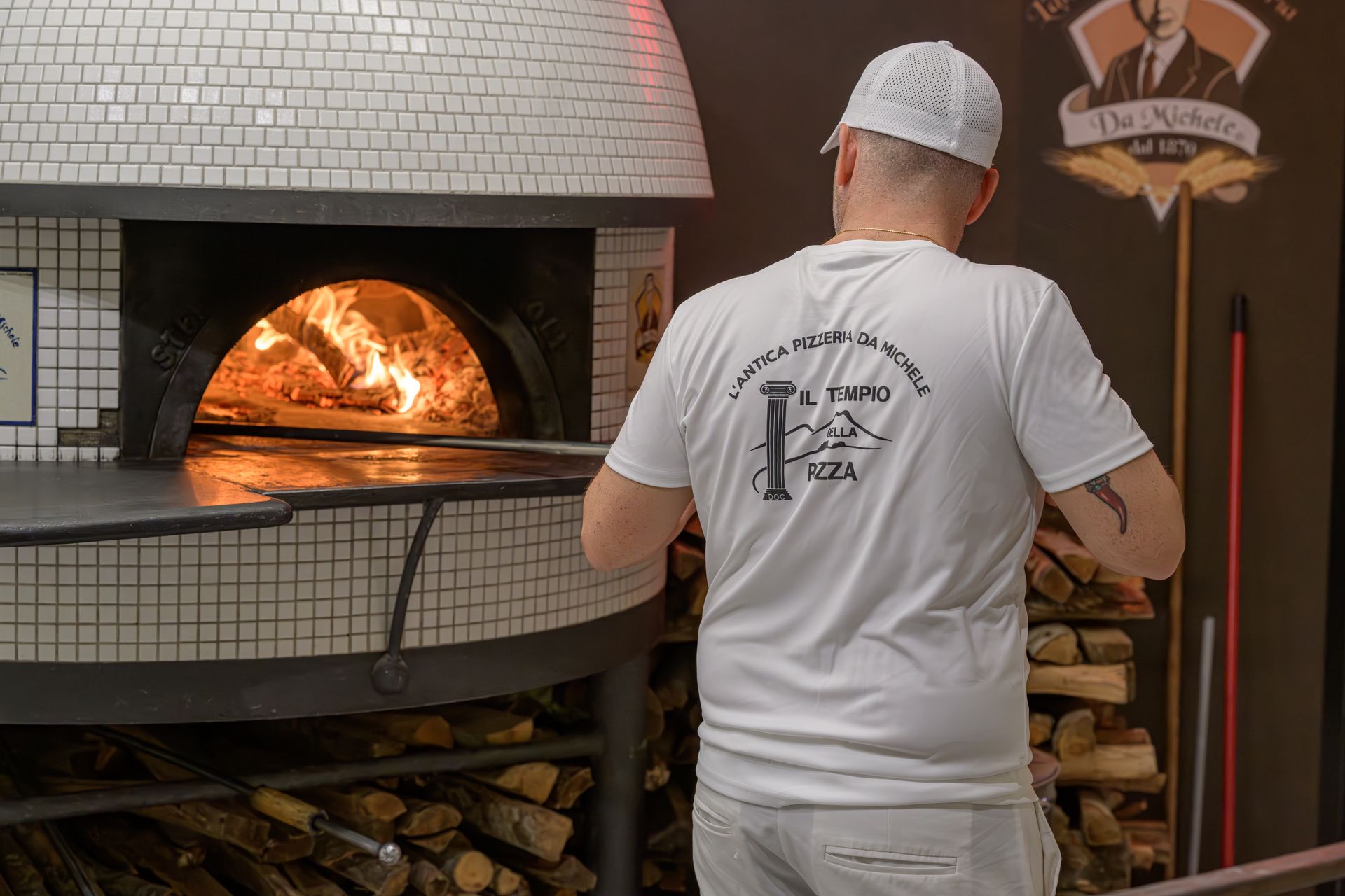 Uomo che si occupa di un forno a legna per pizza, con indosso un cappello bianco e una camicia, con della legna da ardere accatastata lì vicino.