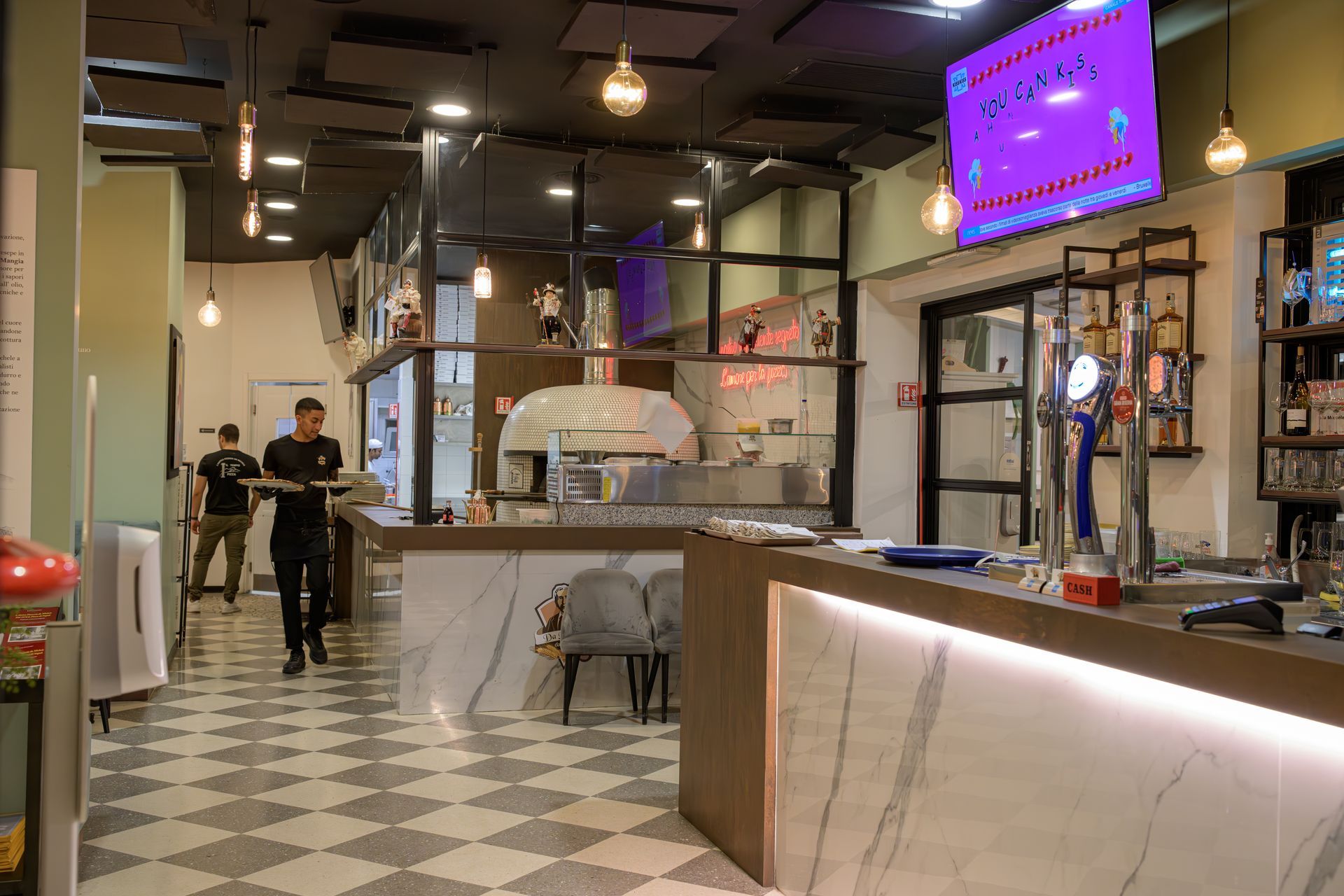 Interno di un ristorante con forno per pizza, bar, pavimento a scacchi e personale che trasporta vassoi.