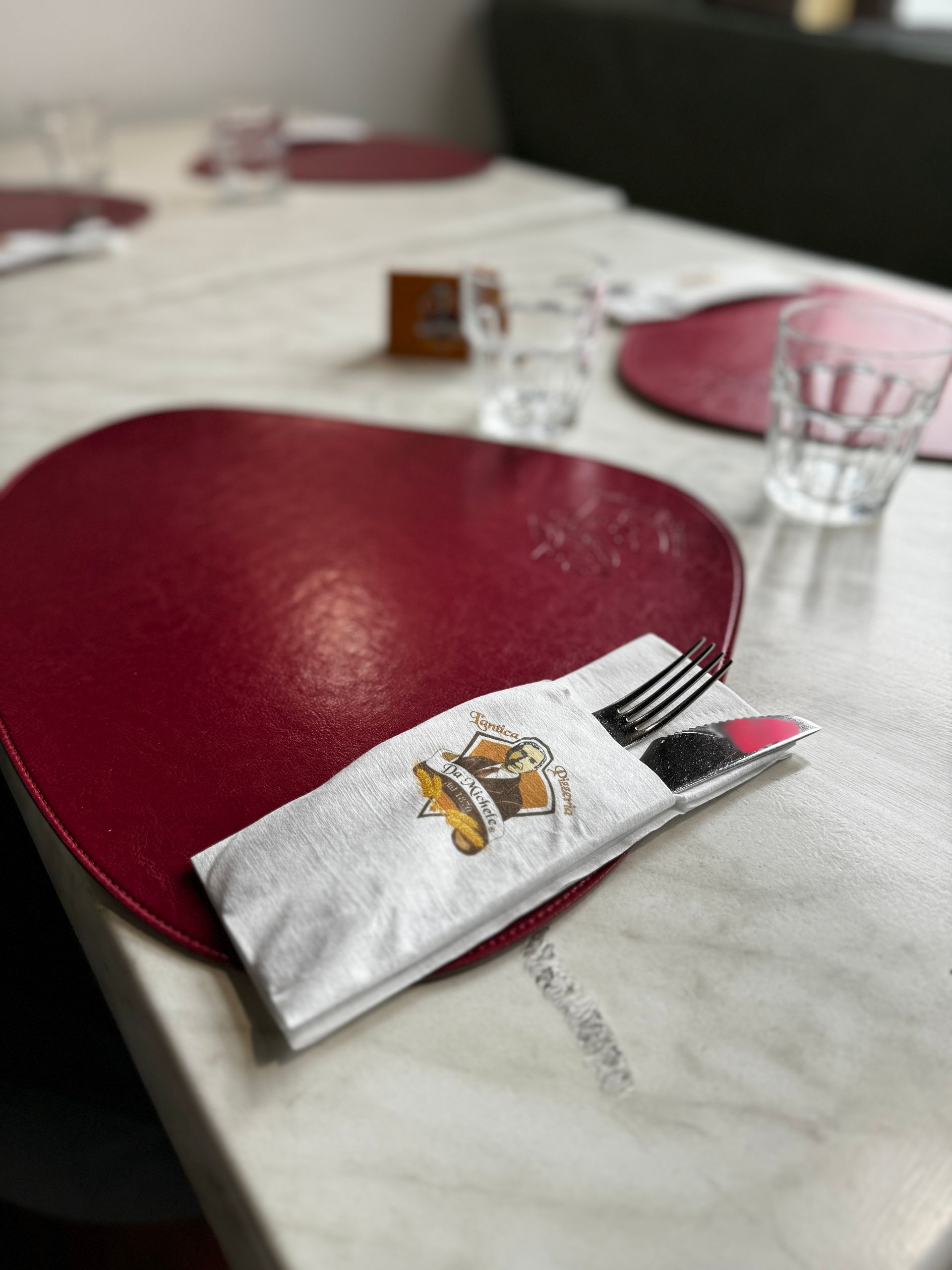 Apparecchiatura della tavola: tovaglietta rossa, posate in un tovagliolo con il logo, bicchieri e un ceppo di legno su un tavolo di marmo.