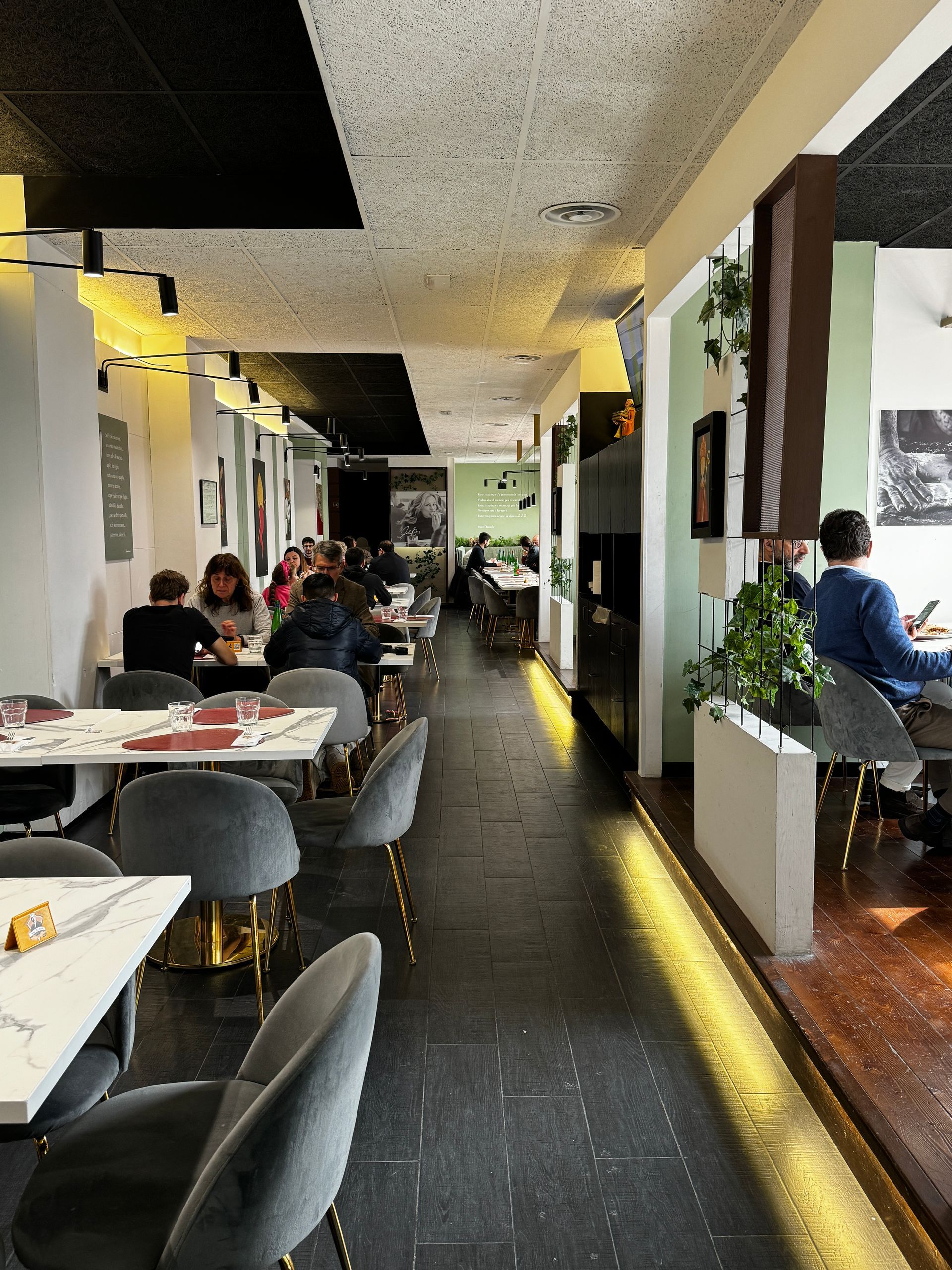 Interno del ristorante: tavoli con commensali, sedie grigie, decorazioni in bianco e nero, piante.
