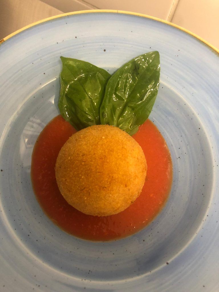 Pallina dorata fritta su salsa rossa, guarnita con foglie di basilico, su un piatto blu.