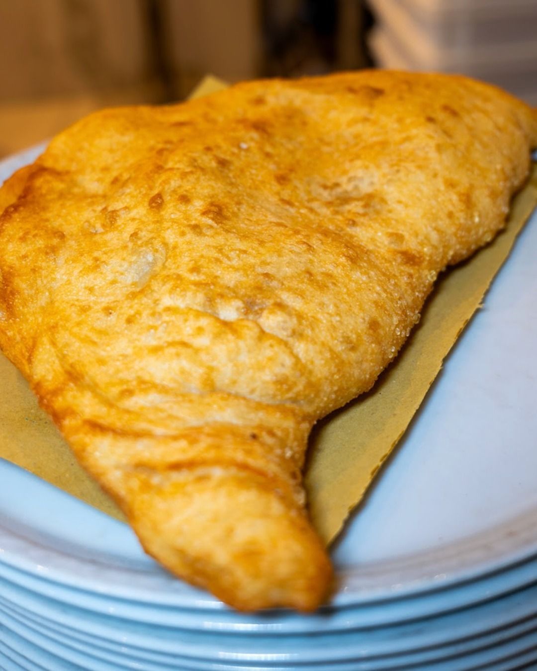 Pasta sfoglia dorata, fritta, simile a una pizza, su un piatto azzurro con una pila sotto; su carta da forno.