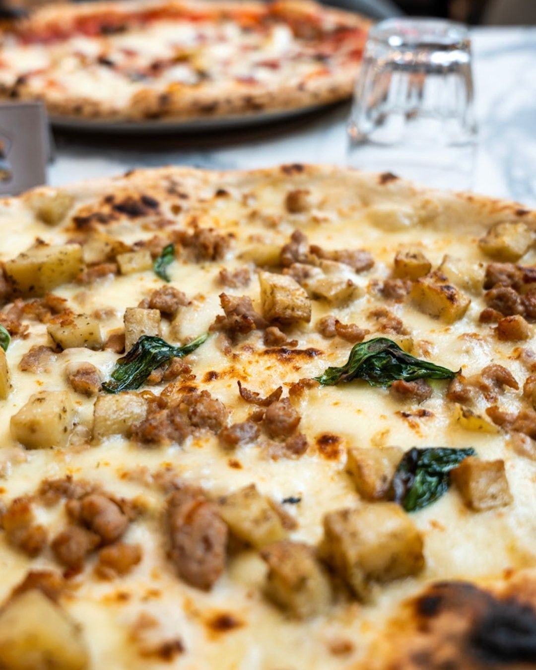 Primo piano di una pizza con salsiccia, patate e basilico. Un'altra pizza è sfocata sullo sfondo.