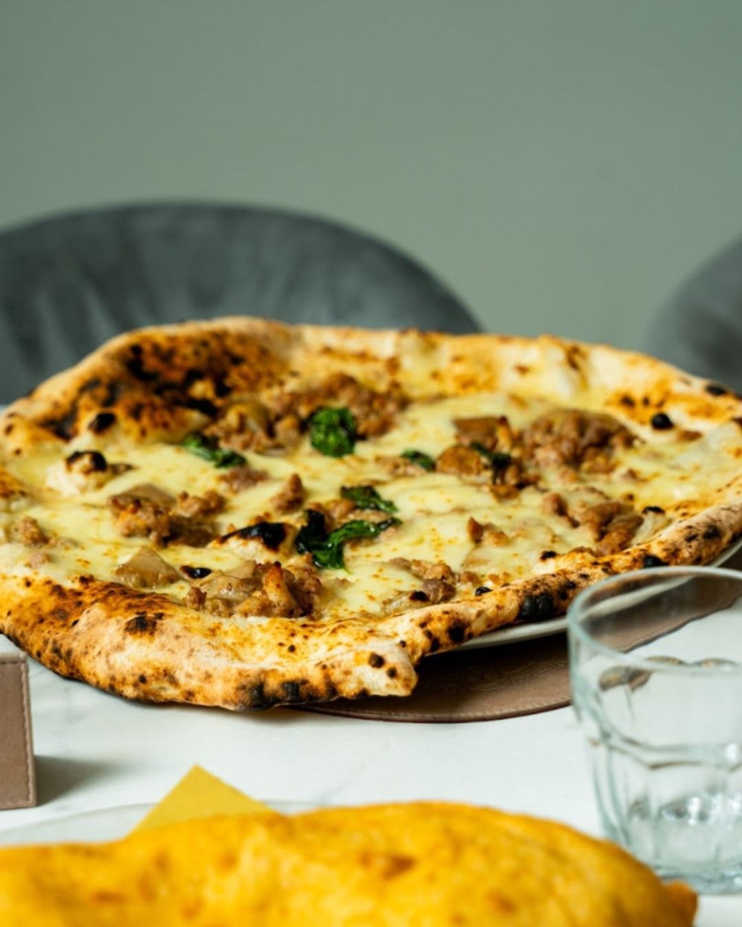 Pizza con condimenti su un tavolo bianco, con un bicchiere d'acqua e una sedia sullo sfondo.