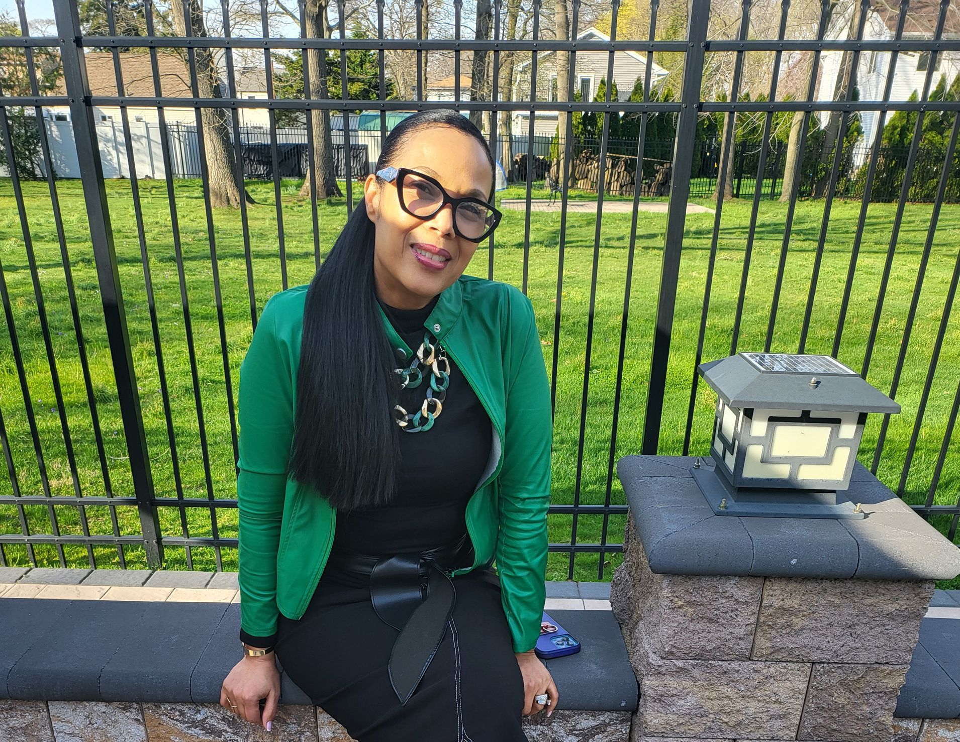 A woman wearing glasses and a green jacket is sitting on a stone wall.