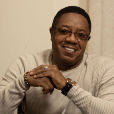 A man wearing glasses and a watch is smiling with his hands folded.