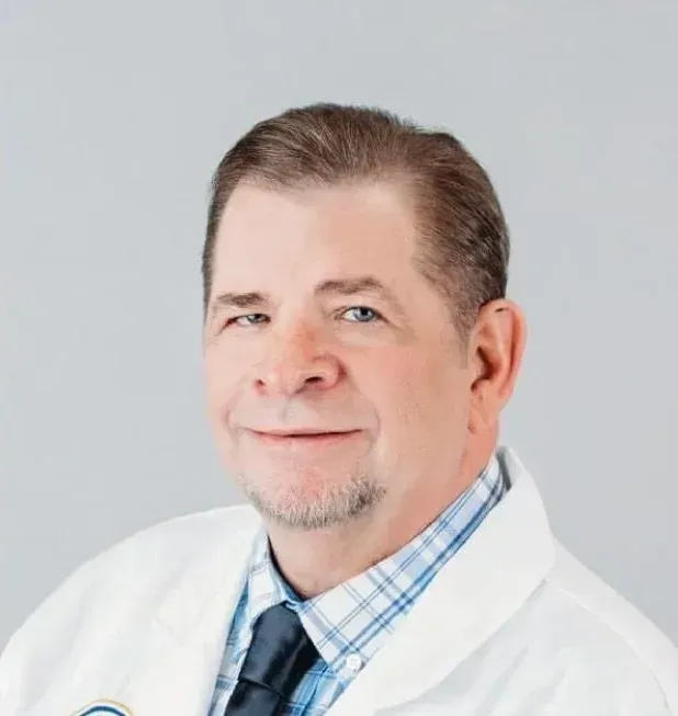 Man in lab coat, smiling, wearing a tie and collared shirt, against a gray background.