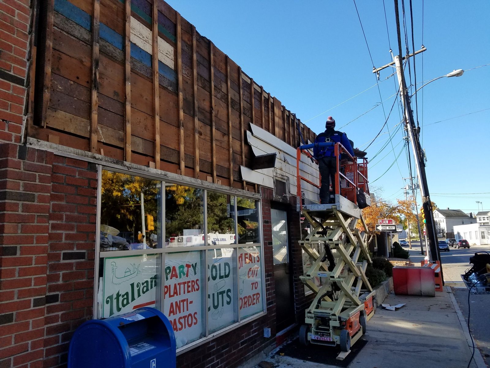 A worker on a lift removes siding from an Italian restaurant building. Brick facade, clear sky, street scene.