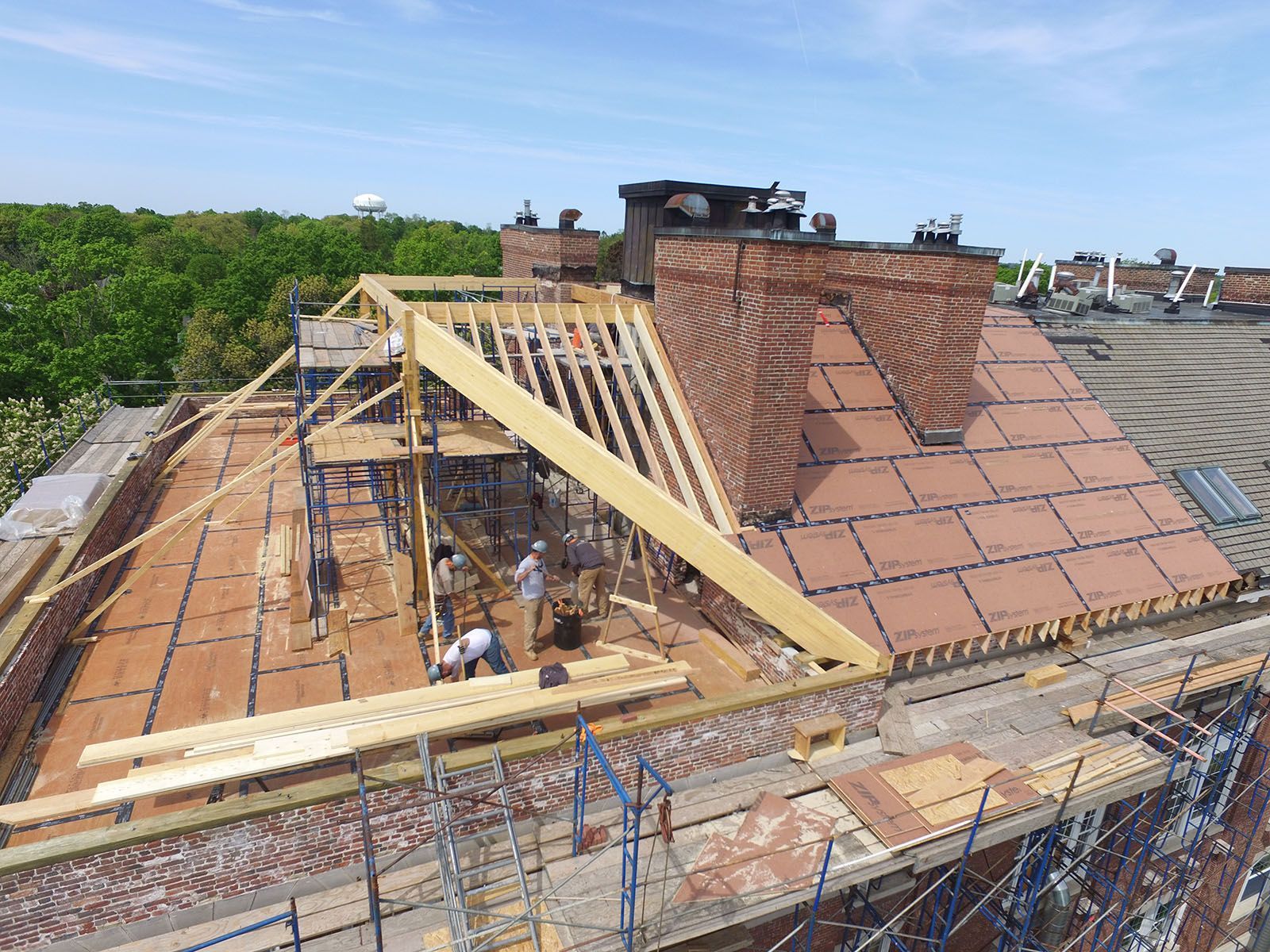 Roof construction with workers, wood beams, brick chimney, and blue sky.