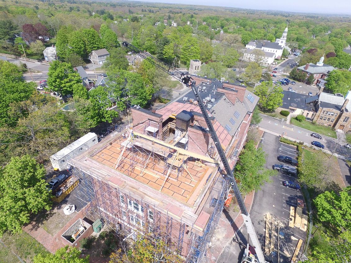 Aerial view of a brick building with roof construction, surrounded by trees and a town.