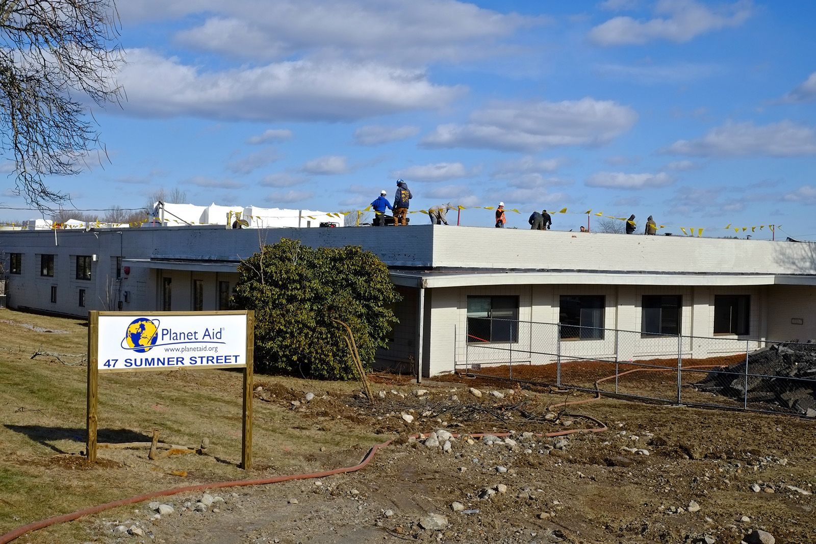 Workers on a flat rooftop of a building, a sign in the foreground, blue sky in the background.