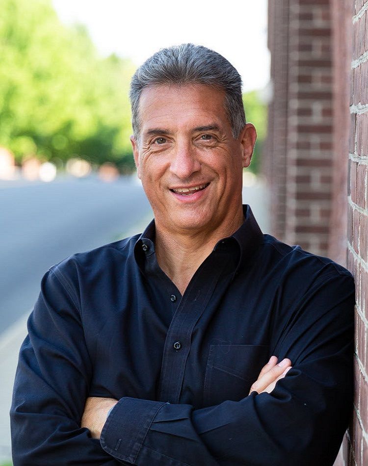 Man in black shirt smiles, arms crossed, leans against a brick wall on a street.